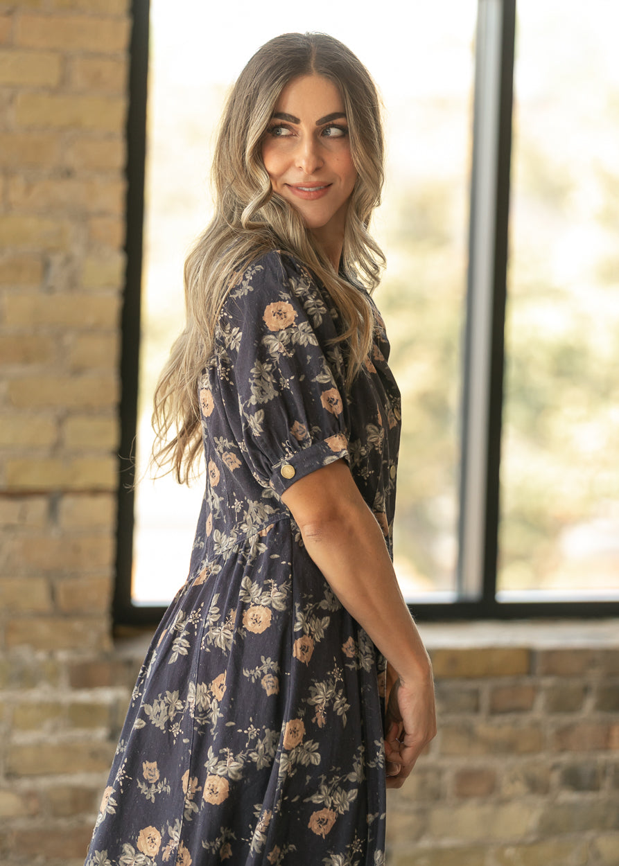 A woman with long wavy blonde hair wearing a dark navy blue floral midi dress, standing in front of a brick wall and a window.