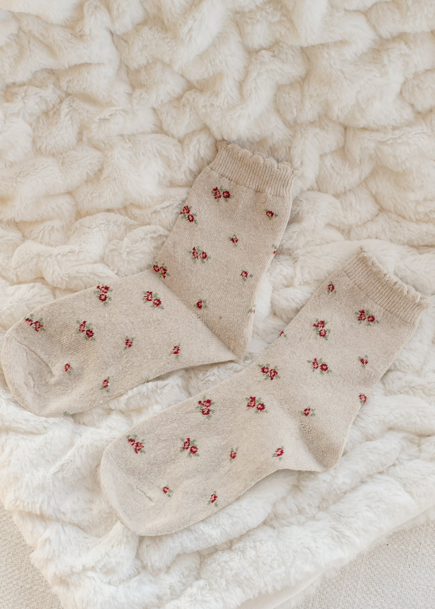 Tan cotton crew socks with delicate red and green floral print and ribbed cuffs, displayed on a cream textured blanket.