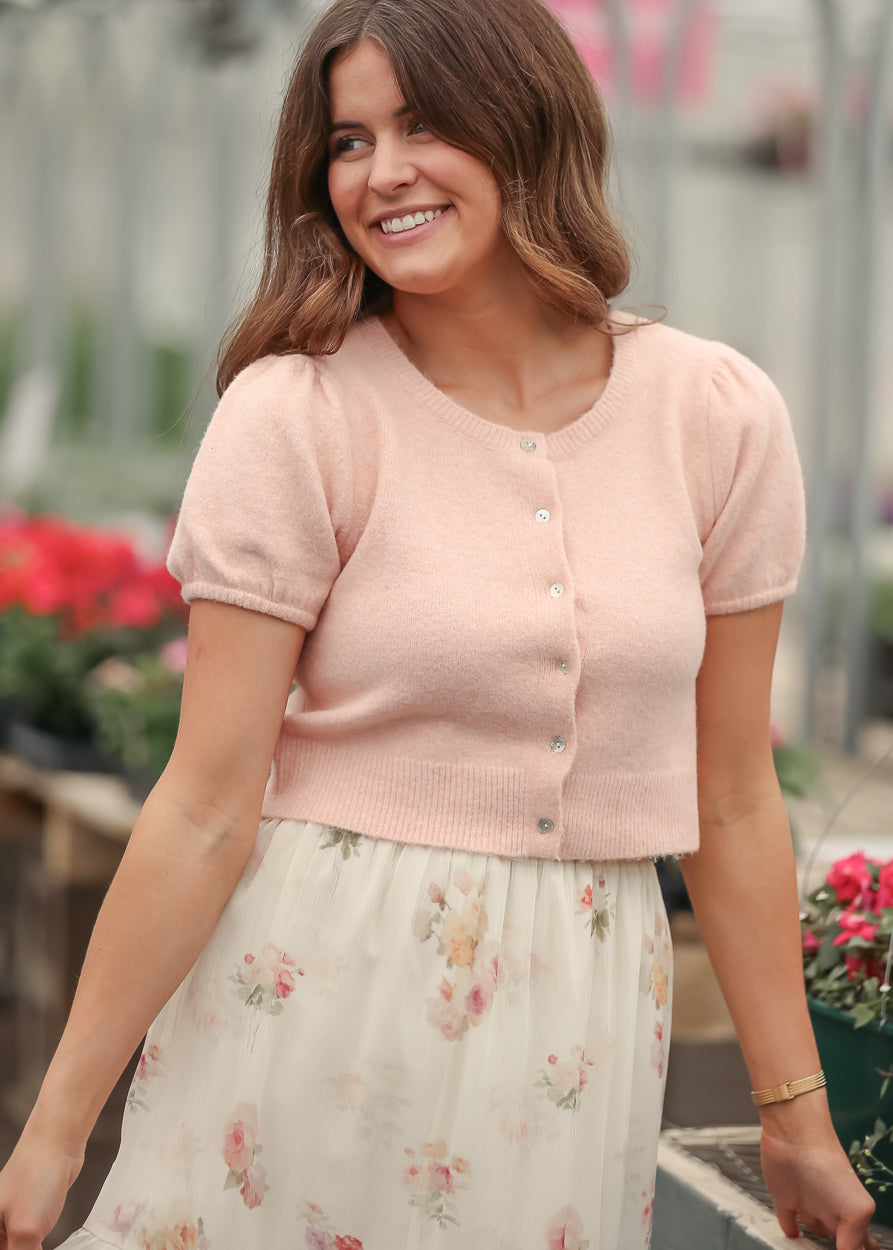 Soft pink cropped button front cardigan with puff sleeves, styled with a floral cream tiered maxi skirt and sandals.