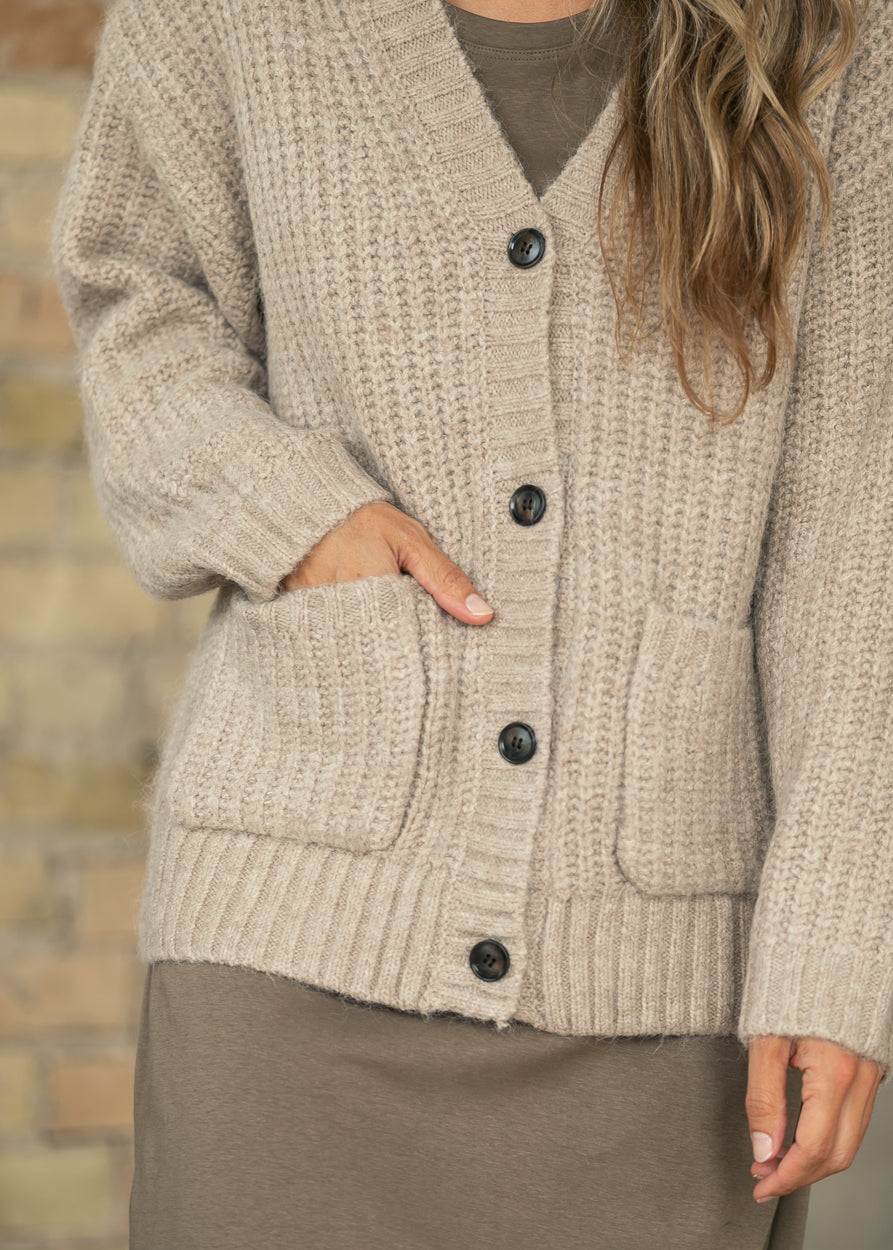 A close-up of a person wearing a chunky knit cardigan in a warm taupe-beige color. The cardigan features a V-neck, dark buttons, and front patch pockets, layered over a matching taupe dress or sweater.