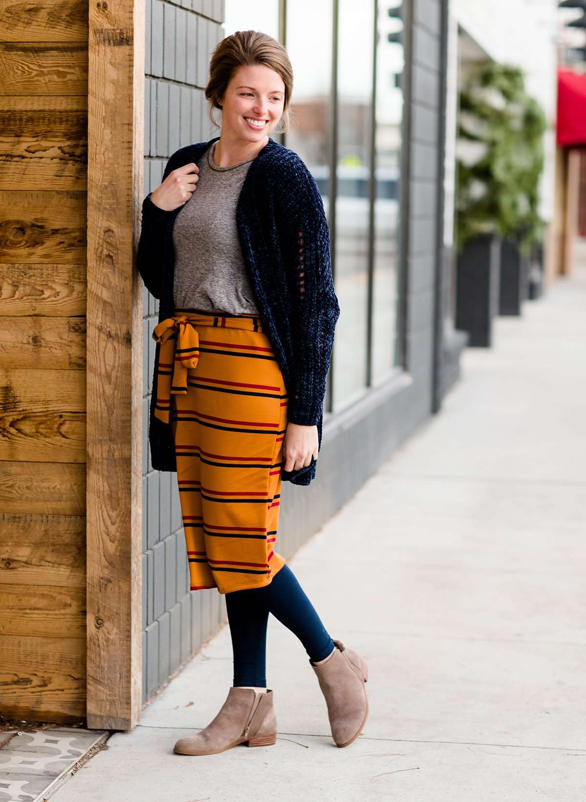 Woman wearing a modest mustard and burgandy striped midi skirt. This skirt has a fabric belt that is removable and is also mommy and me styled. This skirt is paired with tights and boots.