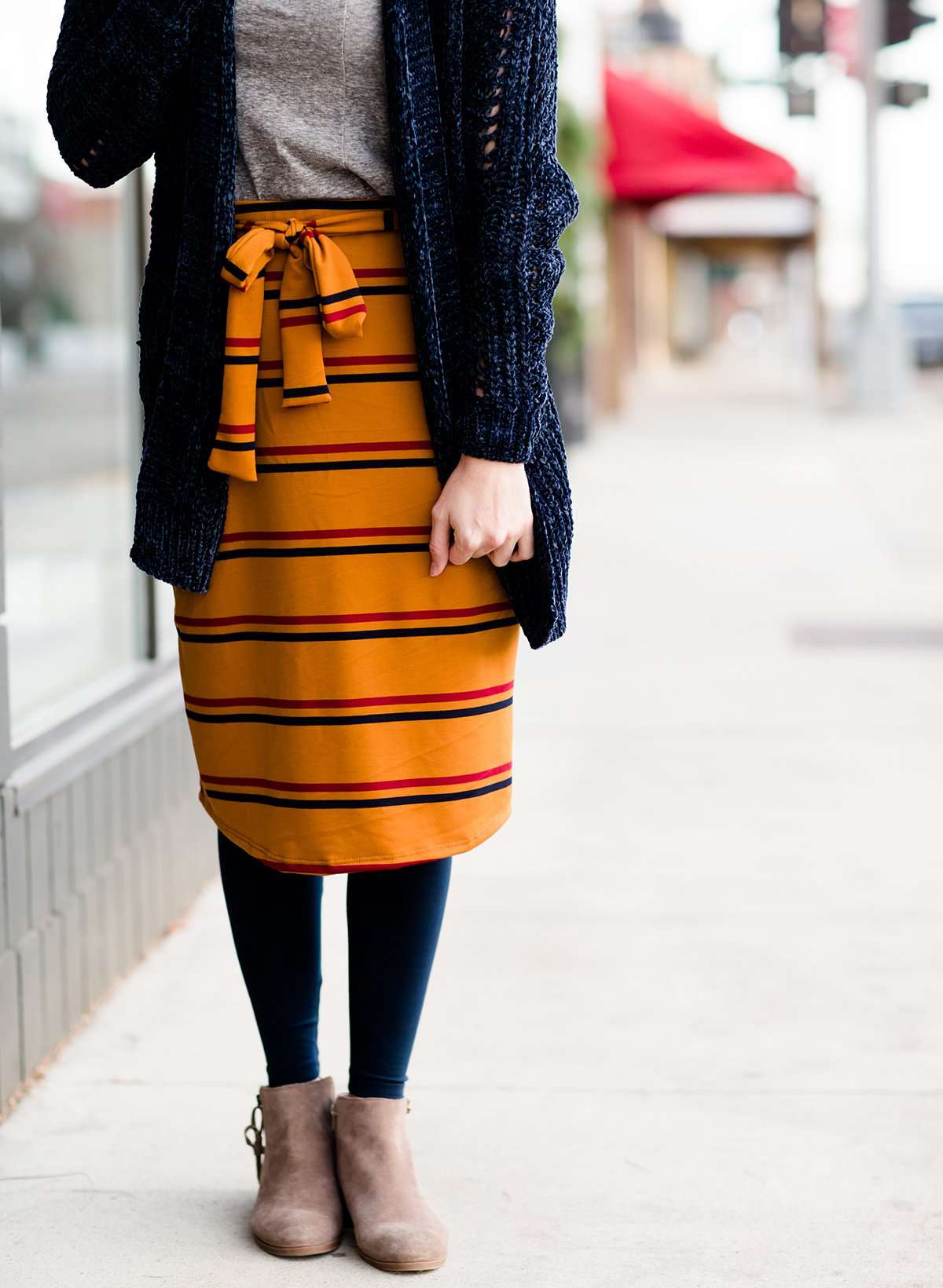 Woman wearing a modest mustard and burgandy striped midi skirt. This skirt has a fabric belt that is removable and is also mommy and me styled. This skirt is paired with tights and boots.