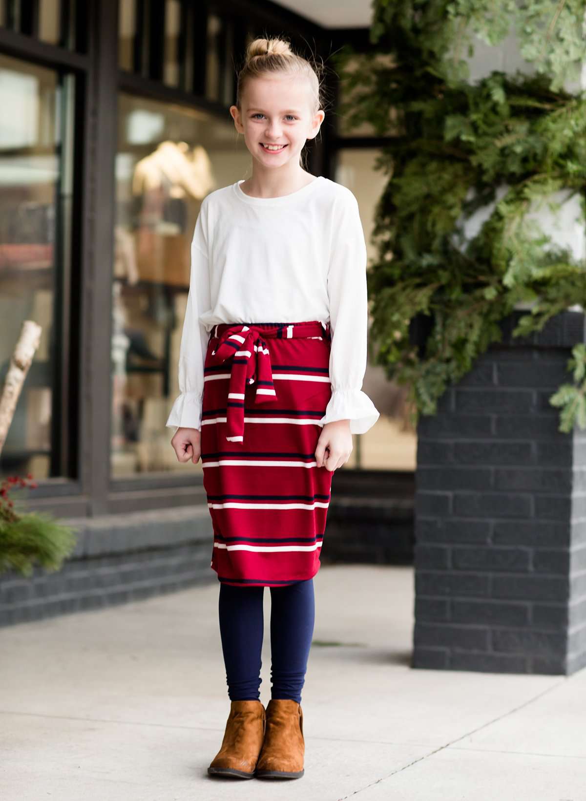 Young girl wearing a burgandy and mustard striped midi skirt with removable belt. This is also a mommy and me skirt. It is paired with a white ruffle top, leggings and brown boots.