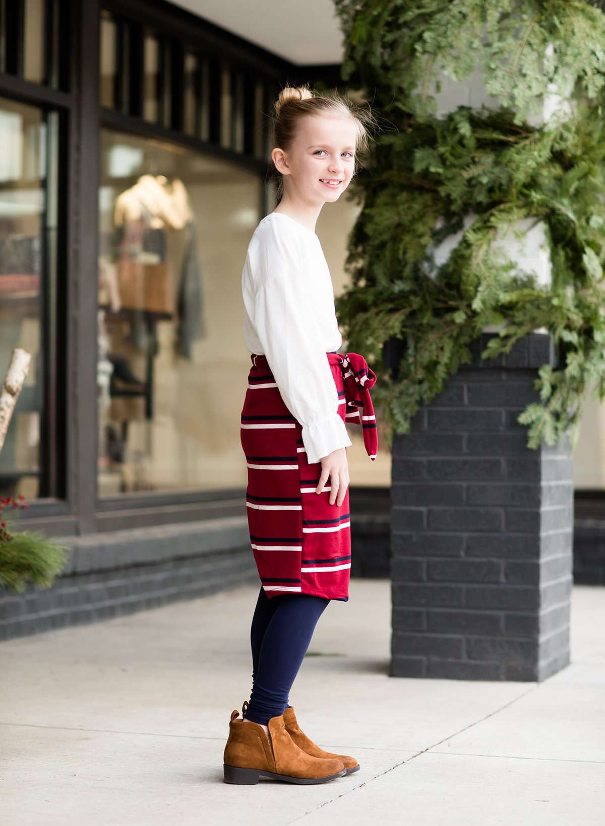 Young girl wearing a burgandy and mustard striped midi skirt with removable belt. This is also a mommy and me skirt. It is paired with a white ruffle top, leggings and brown boots.