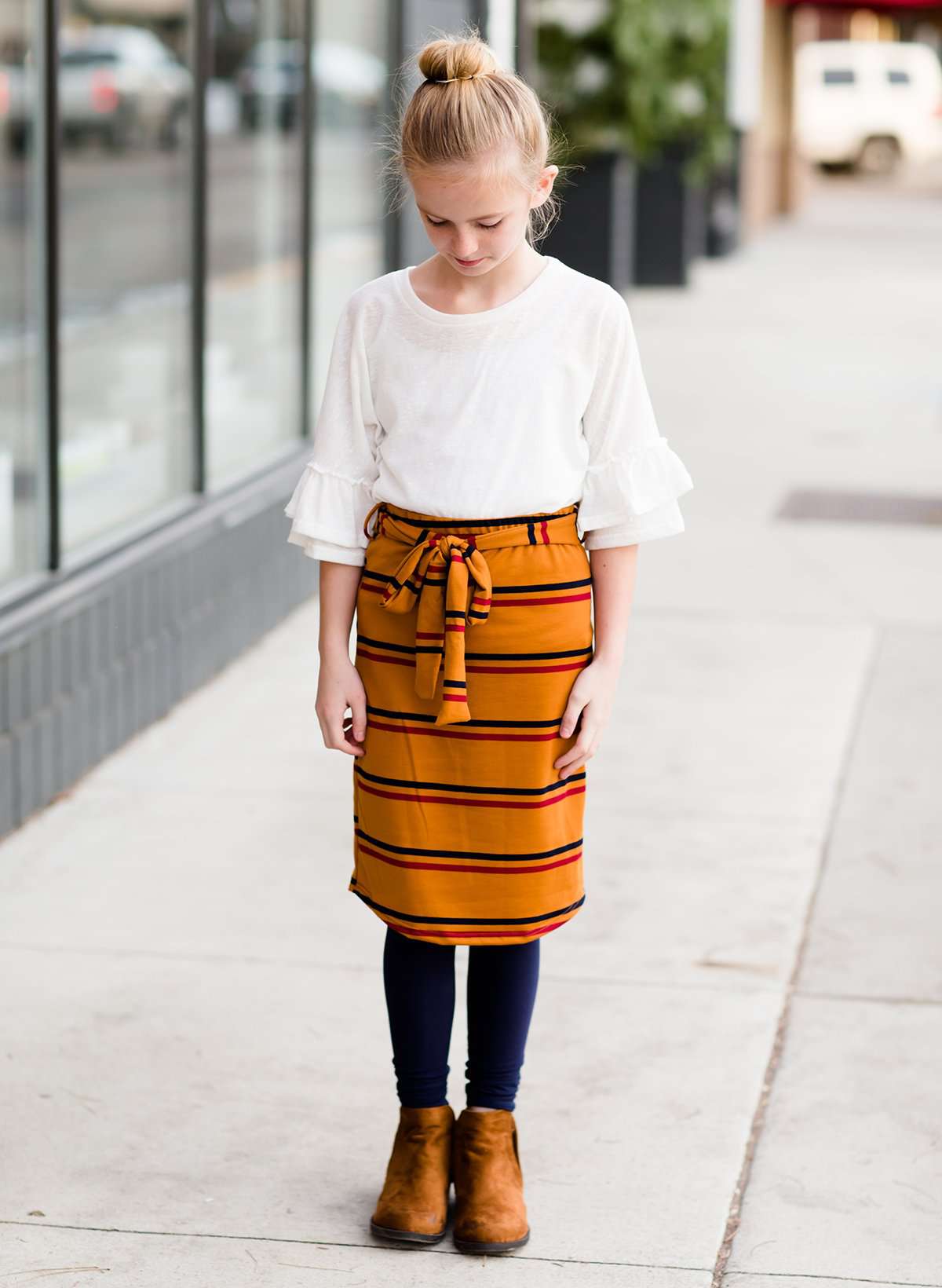 Young girl wearing a burgandy and mustard striped midi skirt with removable belt. This is also a mommy and me skirt. It is paired with a white ruffle top, leggings and brown boots.