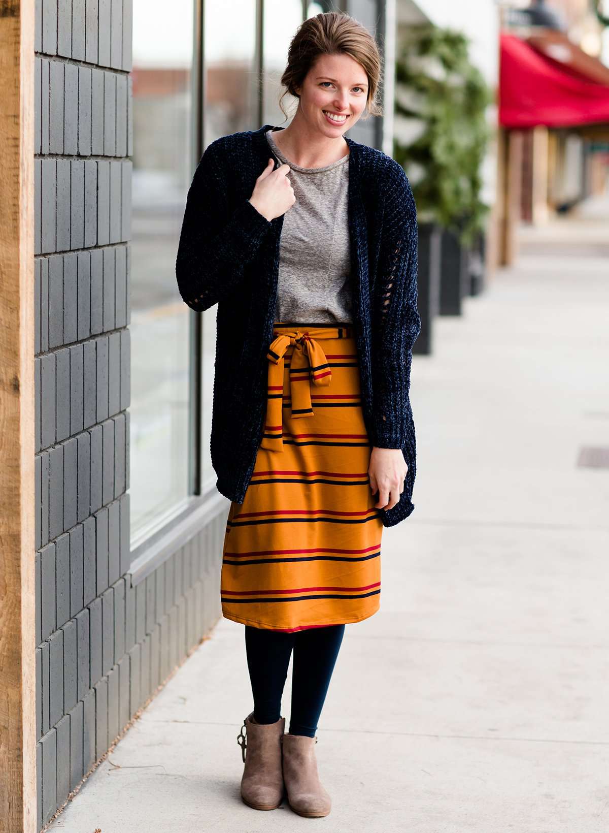 Woman wearing a modest mustard and burgandy striped midi skirt. This skirt has a fabric belt that is removable and is also mommy and me styled. This skirt is paired with tights and boots.