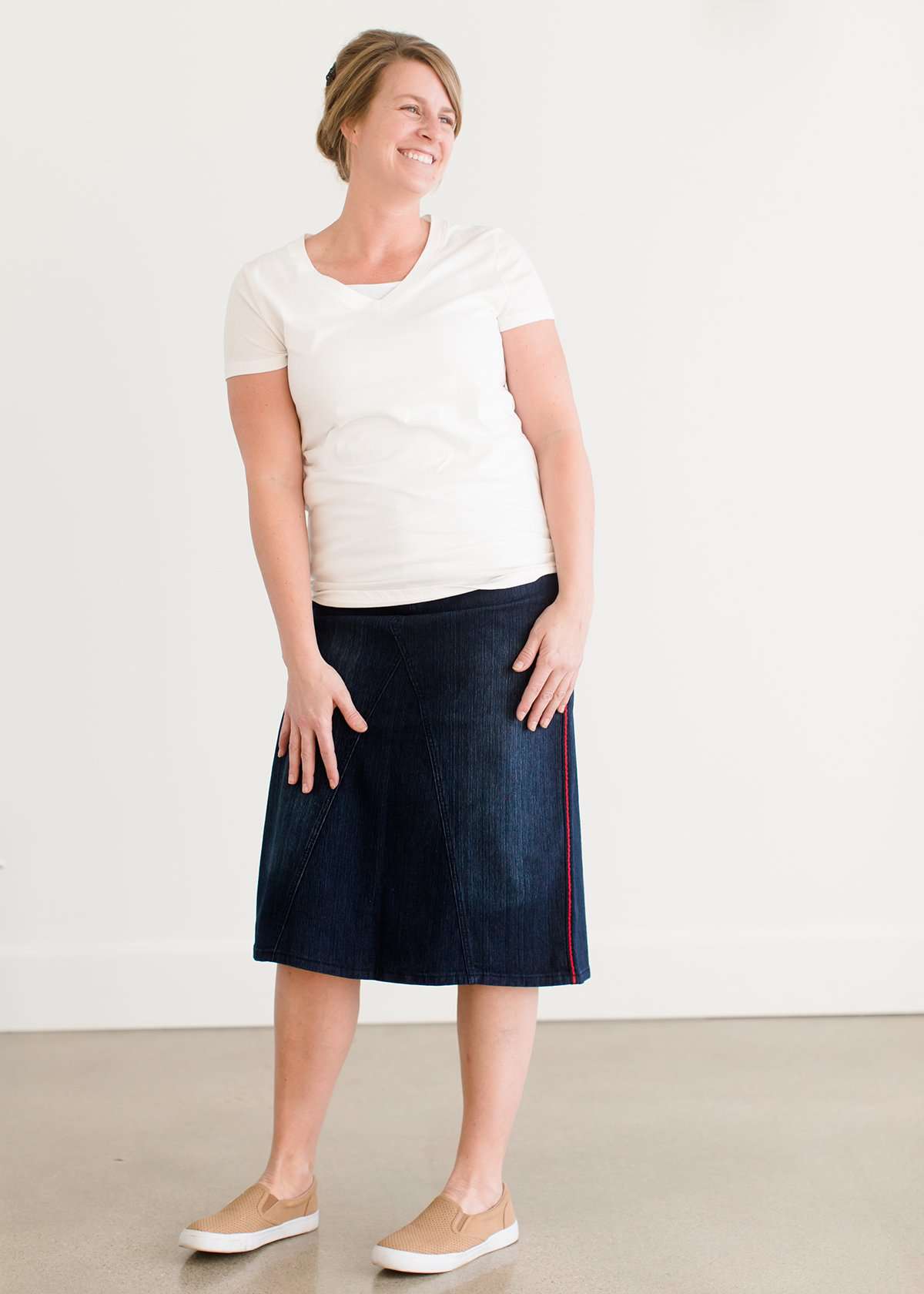 Woman wearing a dark indigo wash below the knee skirt that has red piping along the sides. She also is wearing a white tee shirt and tan sneakers.