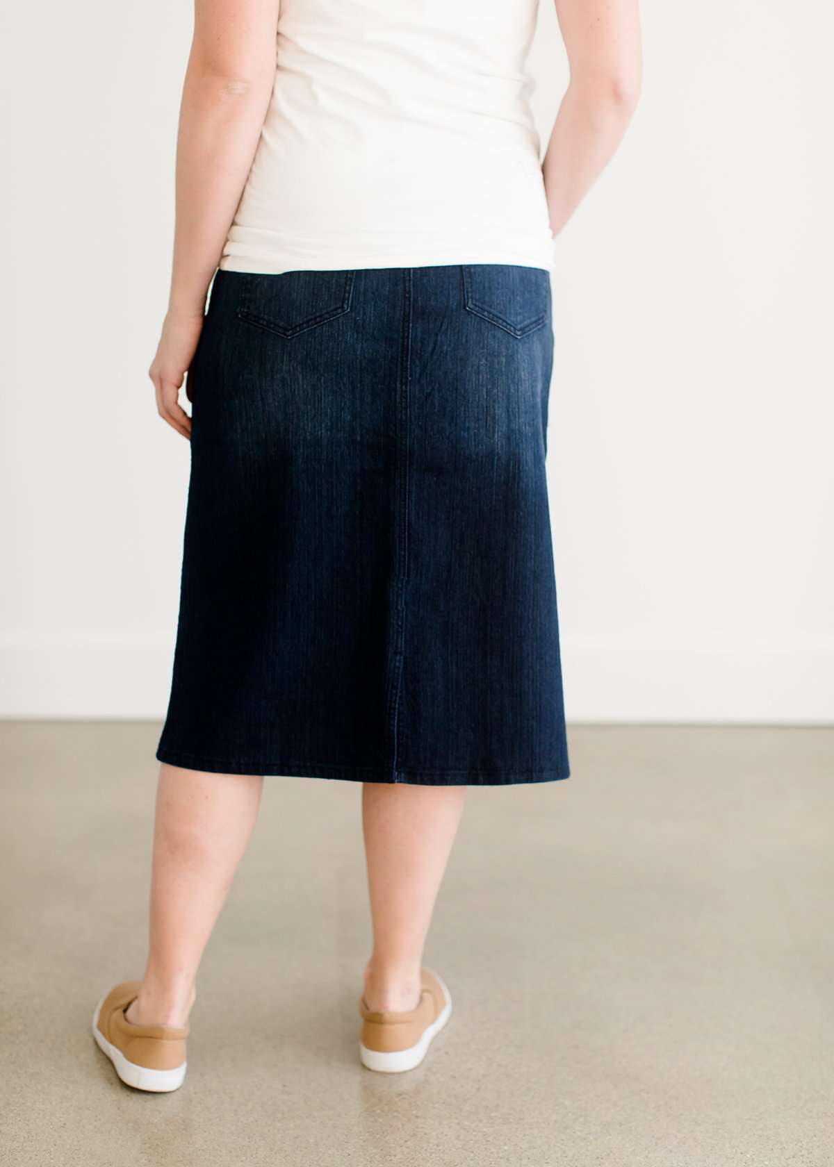 Woman wearing a dark indigo wash below the knee skirt that has red piping along the sides. She also is wearing a white tee shirt and tan sneakers.