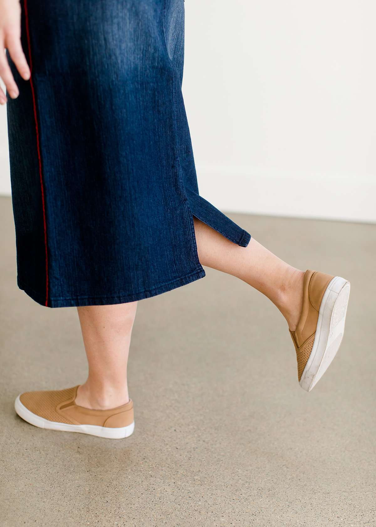 Woman wearing a dark indigo wash below the knee skirt that has red piping along the sides. She also is wearing a white tee shirt and tan sneakers.