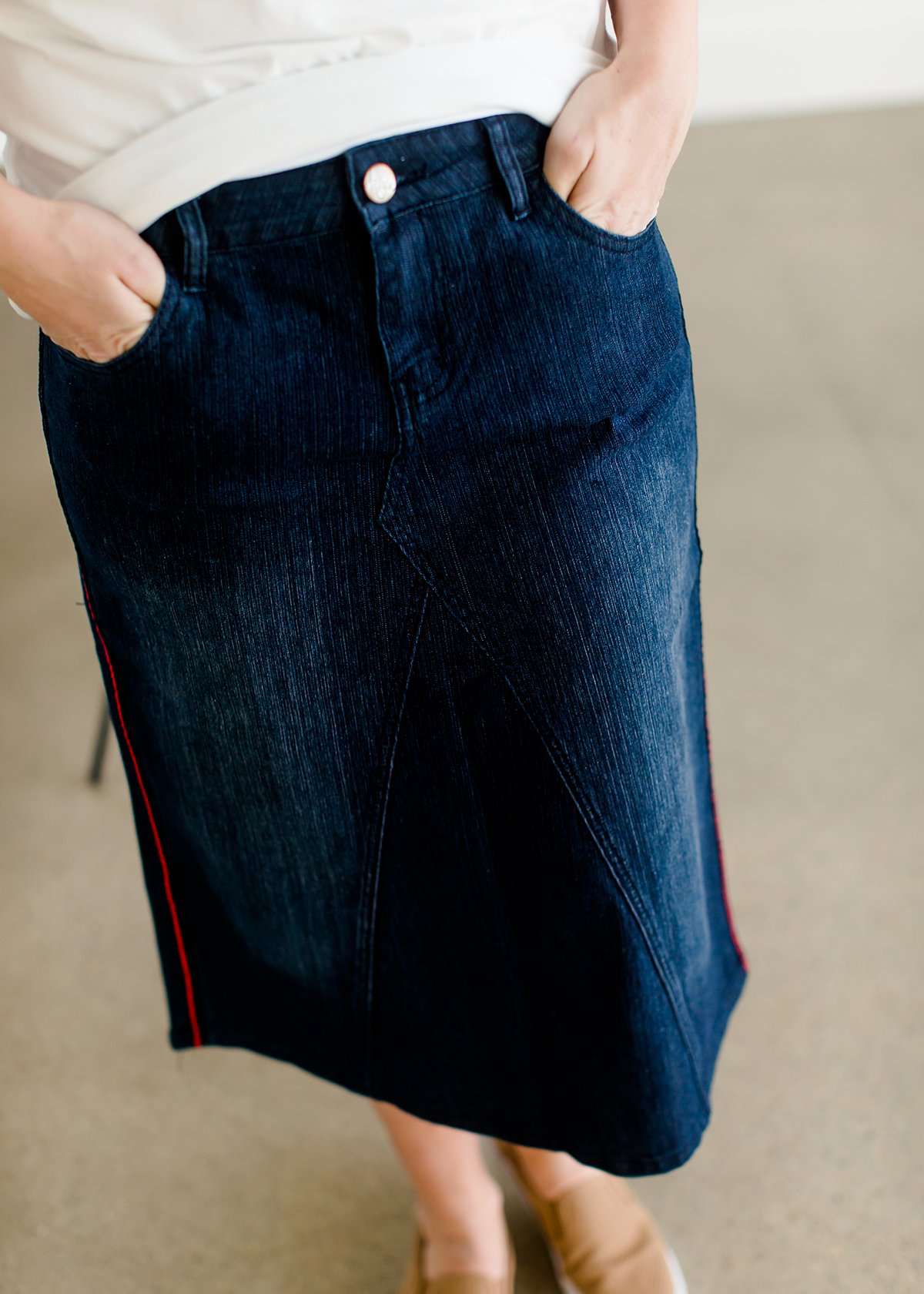 Woman wearing a dark indigo wash below the knee skirt that has red piping along the sides. She also is wearing a white tee shirt and tan sneakers.