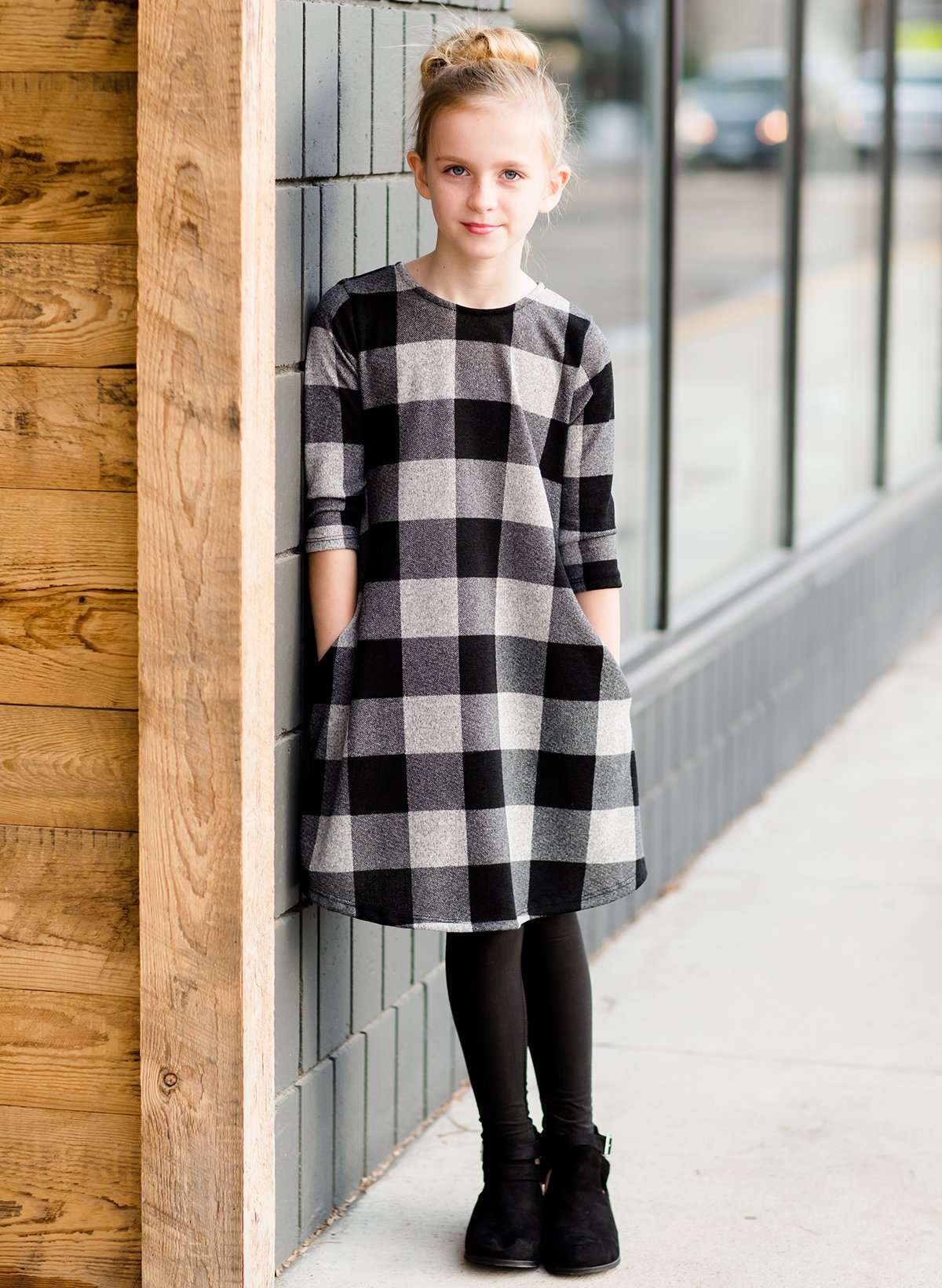 Young girl wearing a black and white buffalo check midi dress. This dress has 3/4 sleeves and front pockets. It is paired with black leggings and black booties.