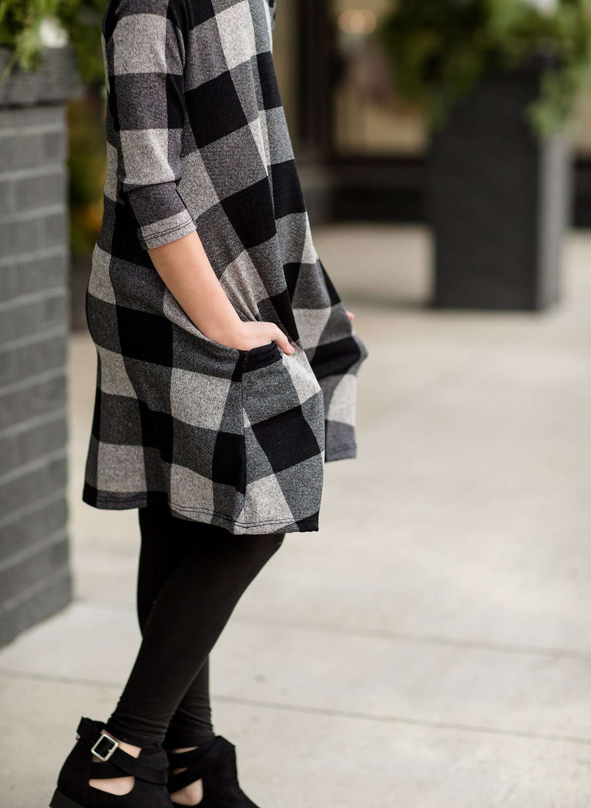 Young girl wearing a black and white buffalo check midi dress. This dress has 3/4 sleeves and front pockets. It is paired with black leggings and black booties.