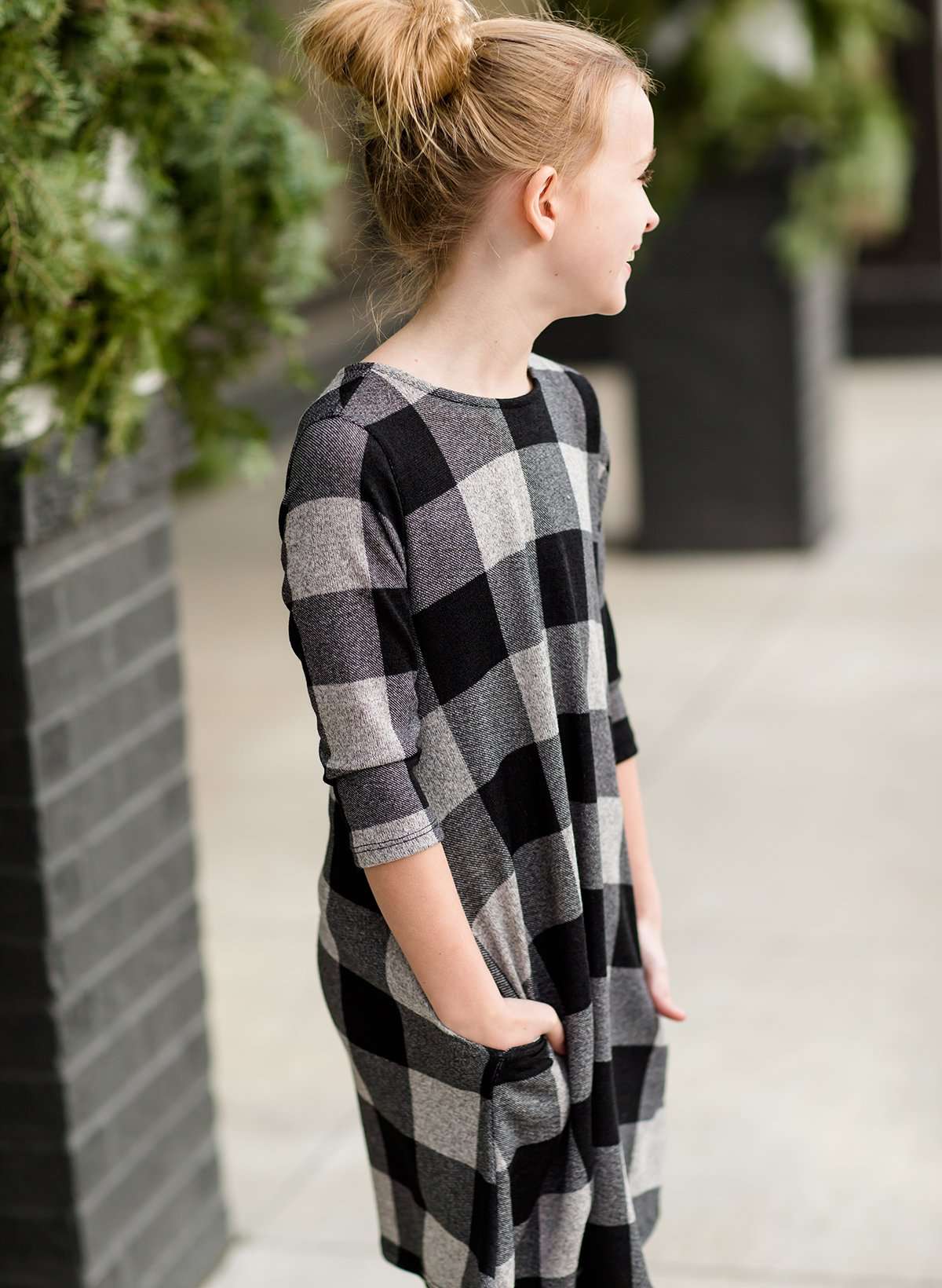 Young girl wearing a black and white buffalo check midi dress. This dress has 3/4 sleeves and front pockets. It is paired with black leggings and black booties.