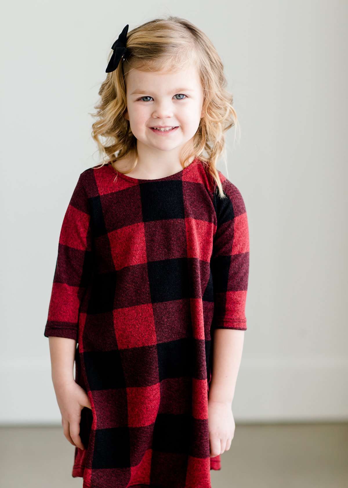 Young girl wearing a black and red buffalo check midi dress. This dress has 3/4 sleeves and front pockets. It is paired with black leggings and black booties.