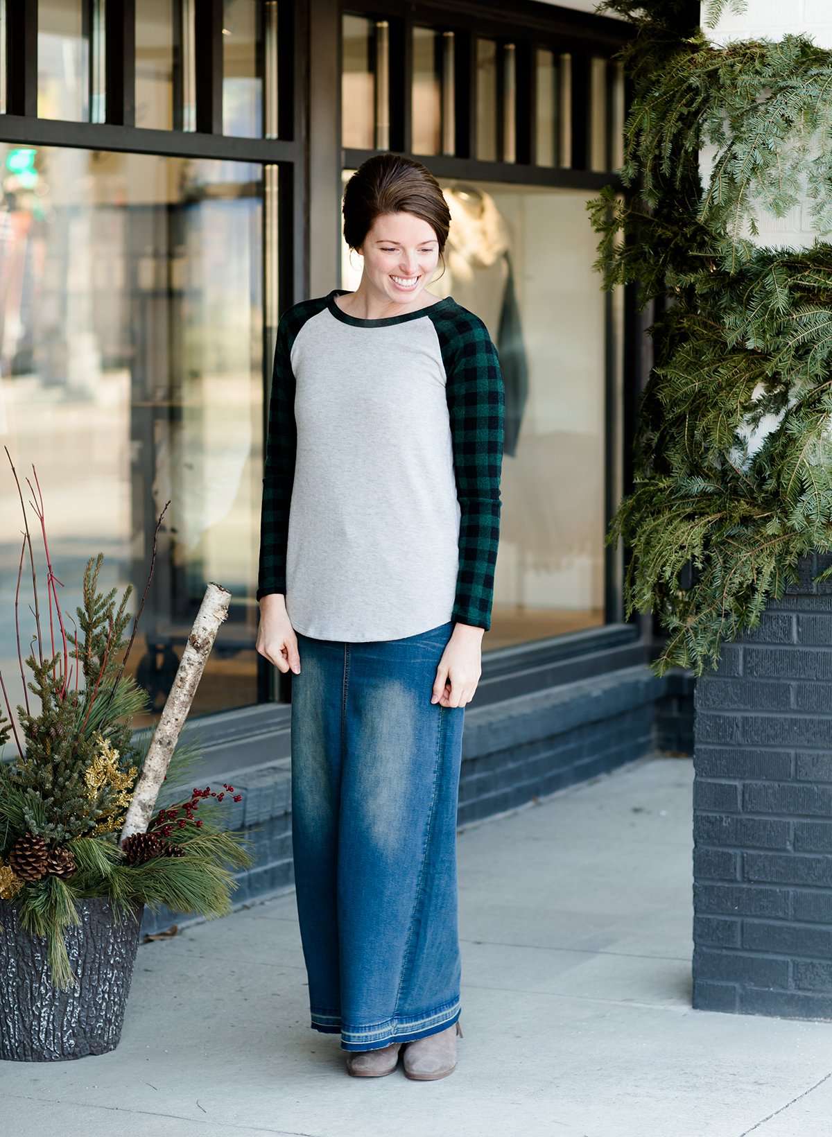 Woman wearing a black and green buffalo check sleeve raglan top paired with a long modest jean skirt standing outside of Inherit Clothing Company!