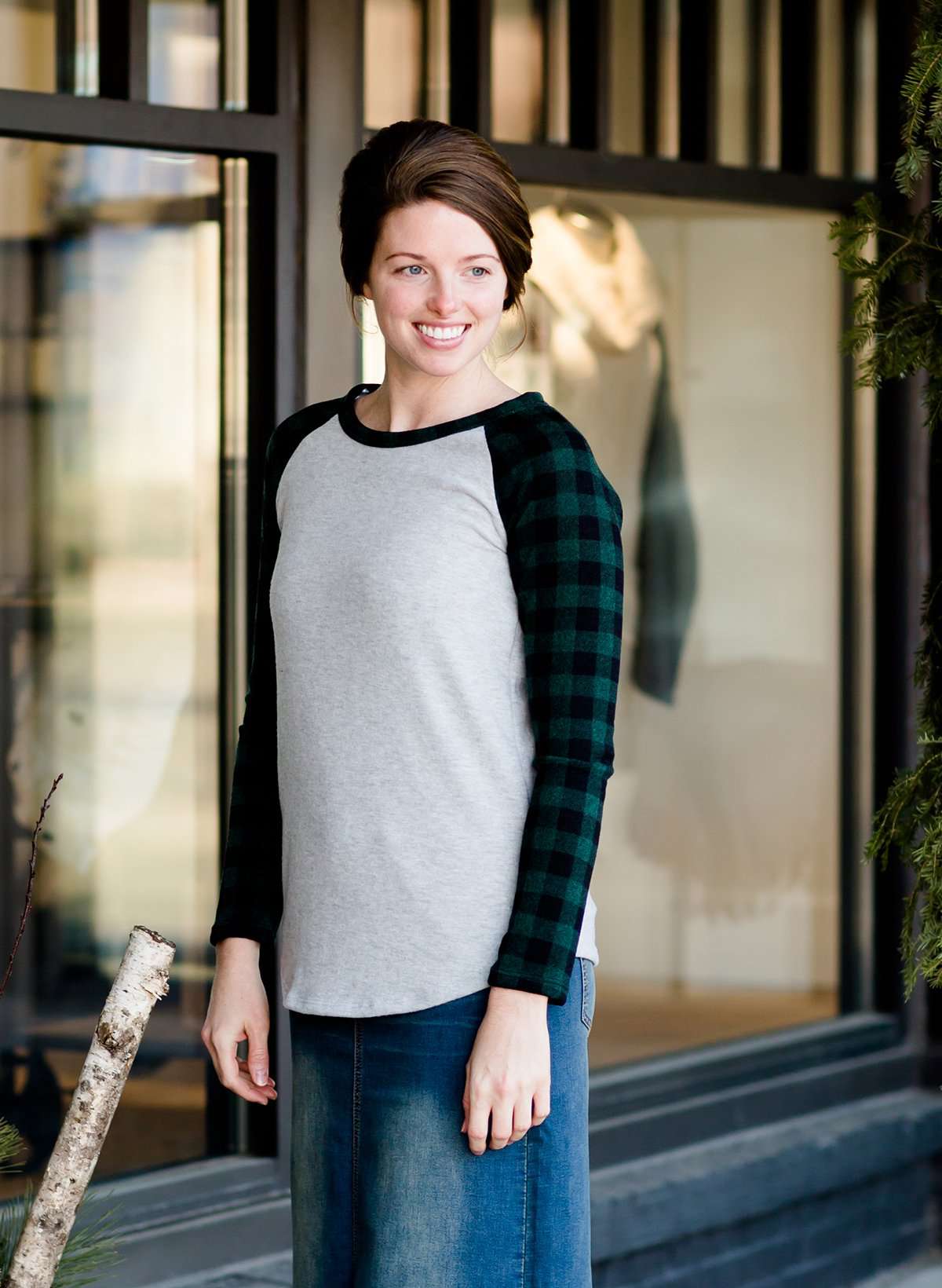 Woman wearing a black and green buffalo check sleeve raglan top paired with a long modest jean skirt standing outside of Inherit Clothing Company!