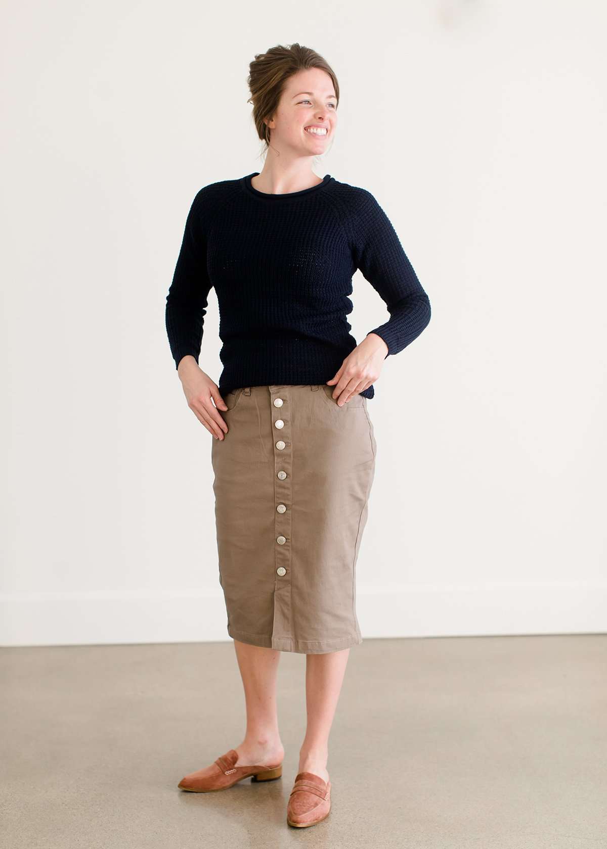 Woman wearing a khaki midi length skirt with faux buttons running down the front. It is paired with a navy sweater and copper, suede slide shoes.