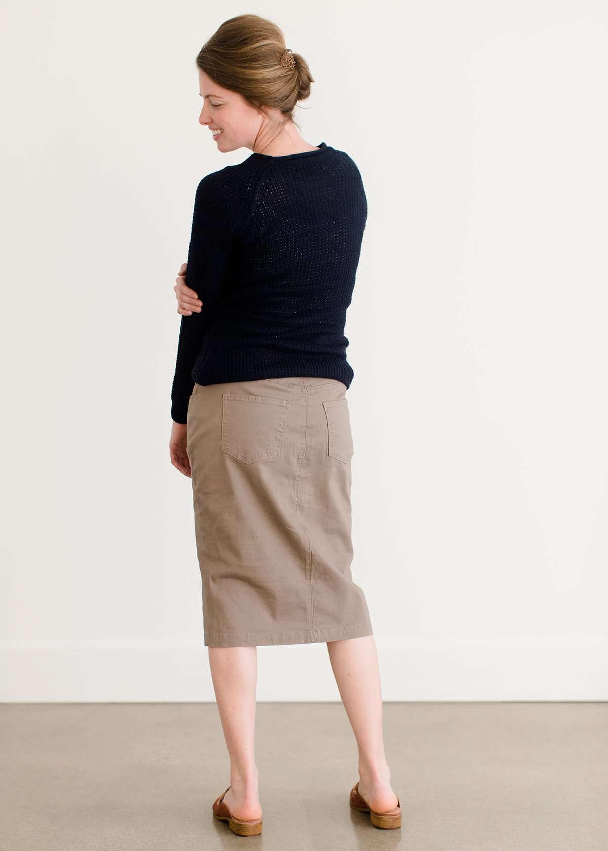 Woman wearing a khaki midi length skirt with faux buttons running down the front. It is paired with a navy sweater and copper, suede slide shoes.