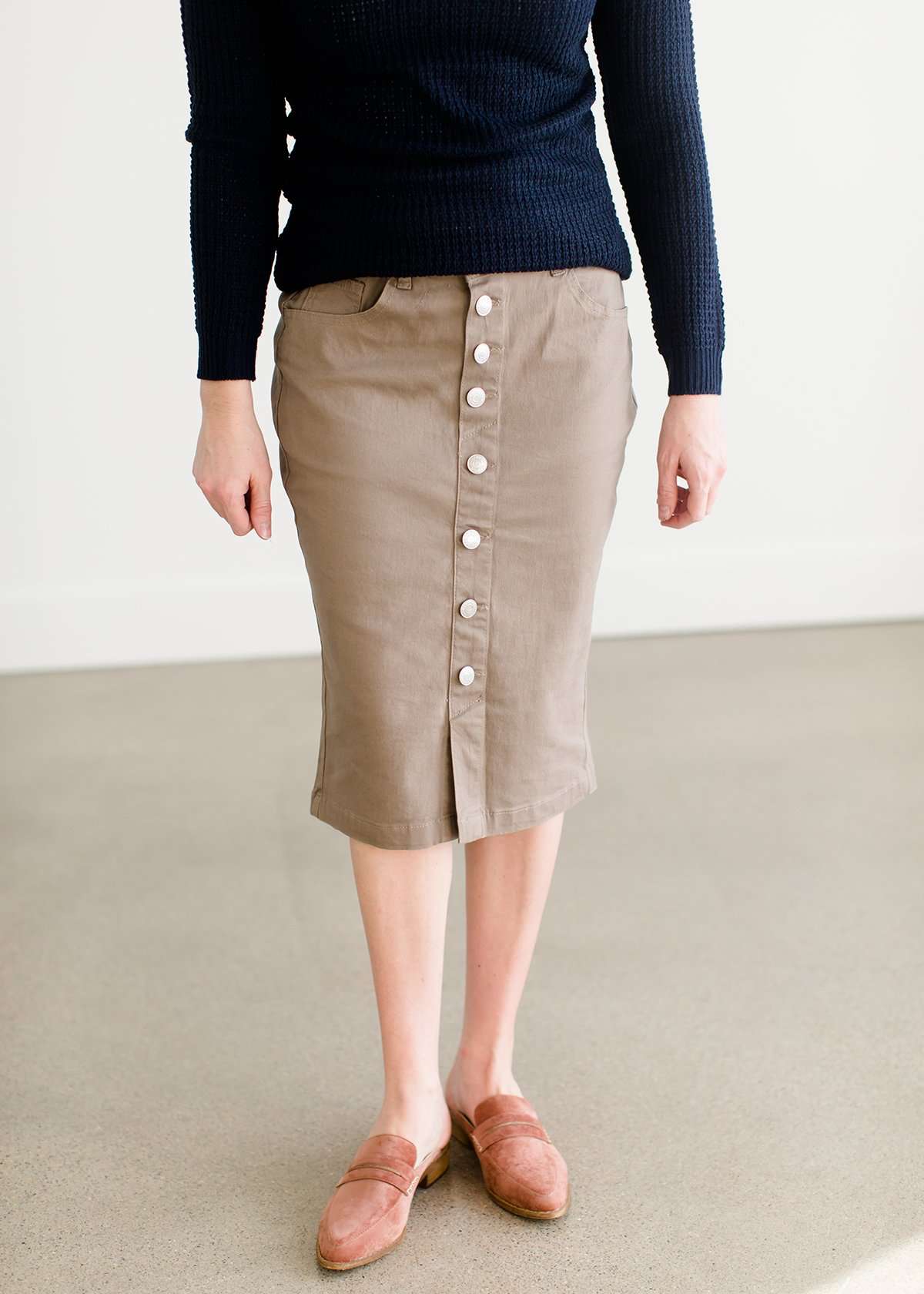 Woman wearing a khaki midi length skirt with faux buttons running down the front. It is paired with a navy sweater and copper, suede slide shoes.