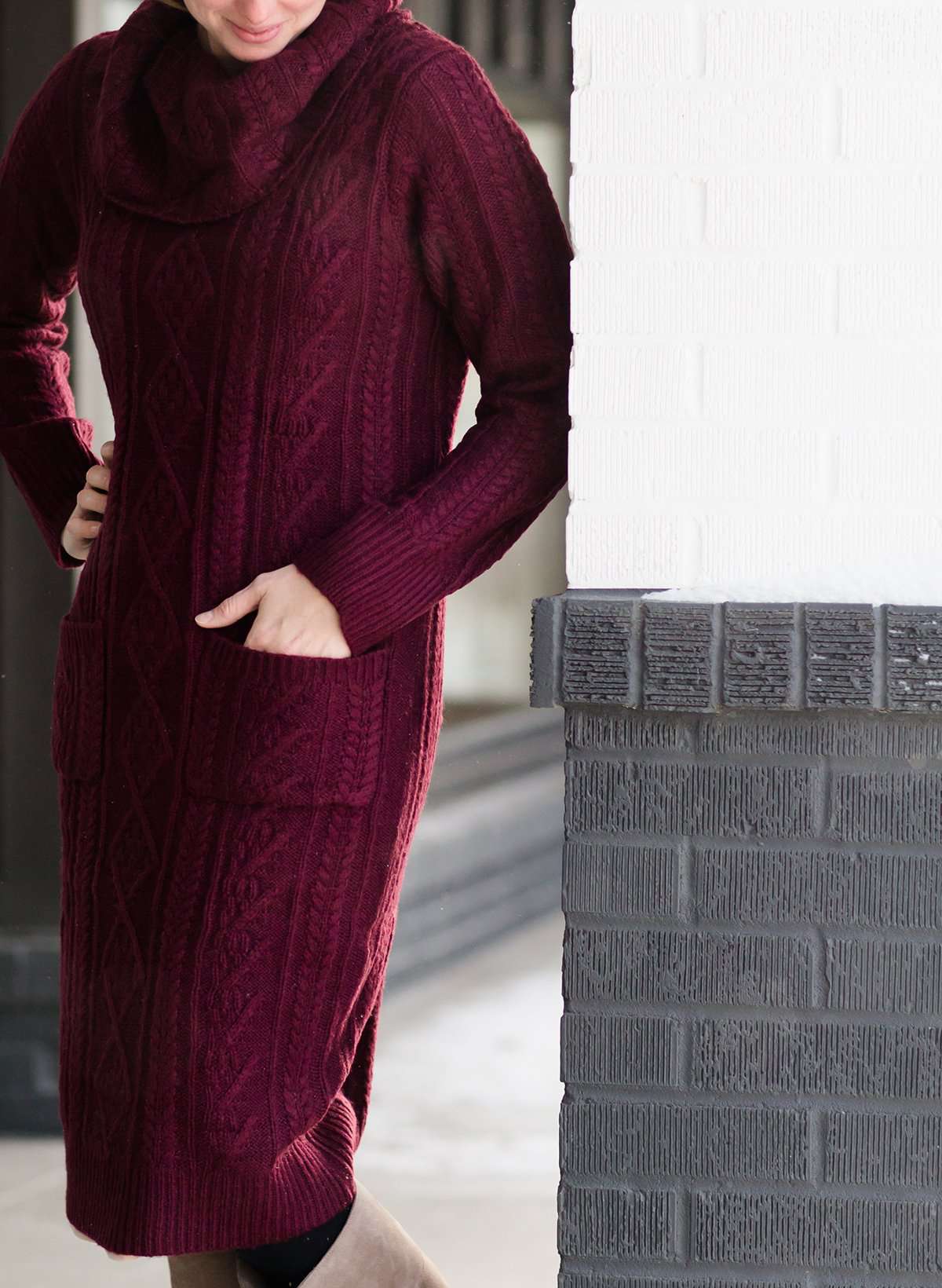 Woman wearing a cableknit, cowlneck sweater dress in taupe or burgandy. This cozy dress is below the knees and paired with tall, suede riding boots.