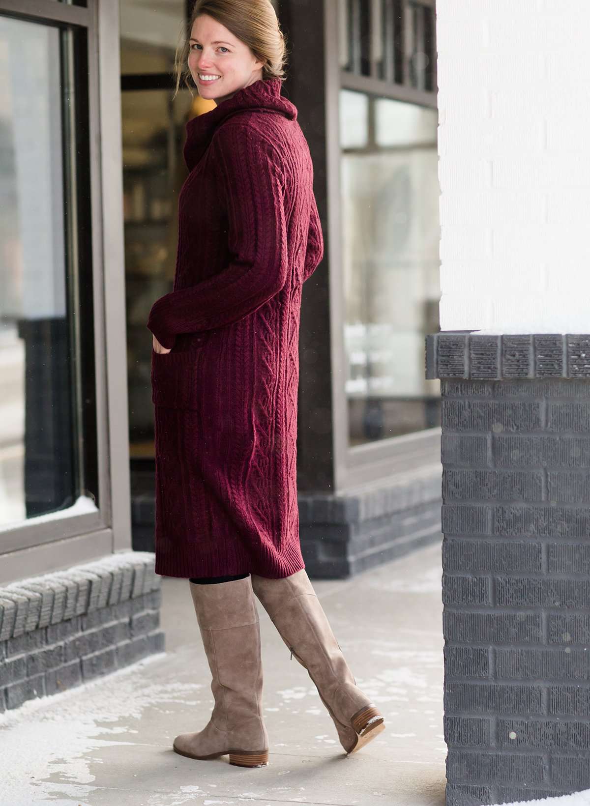 Woman wearing a cableknit, cowlneck sweater dress in taupe or burgandy. This cozy dress is below the knees and paired with tall, suede riding boots.