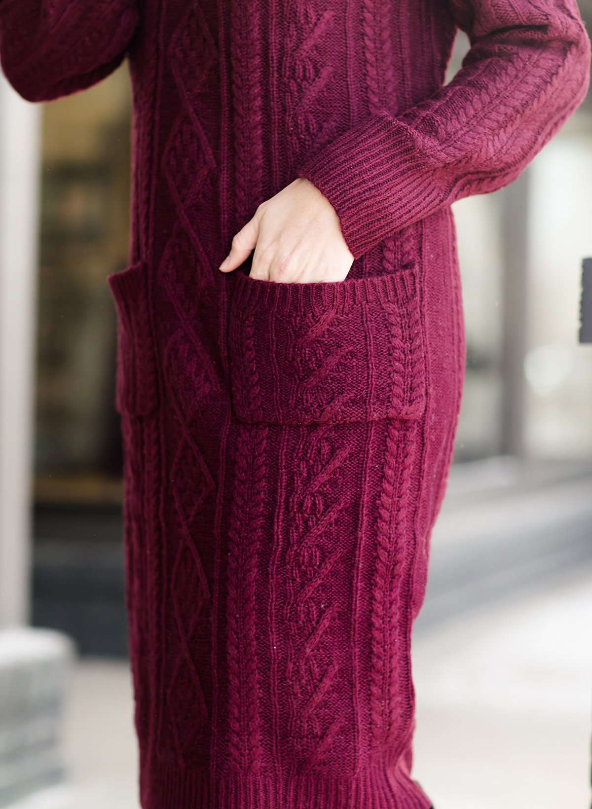 Woman wearing a cableknit, cowlneck sweater dress in taupe or burgandy. This cozy dress is below the knees and paired with tall, suede riding boots.