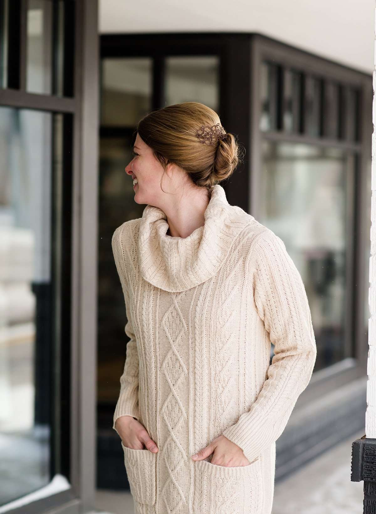 Woman wearing a cableknit, cowlneck sweater dress in taupe or burgandy. This cozy dress is below the knees and paired with tall, suede riding boots.