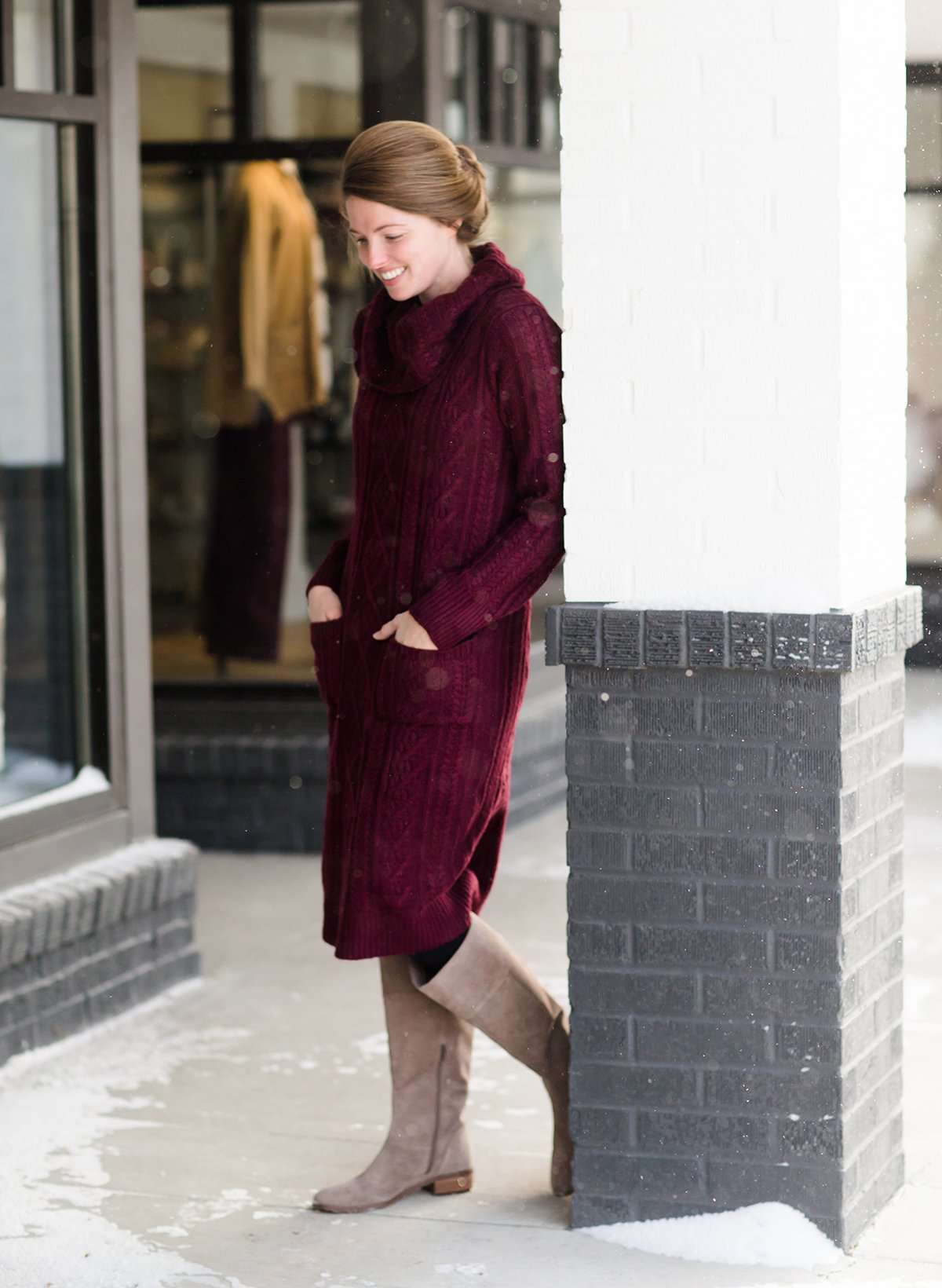 Woman wearing a cableknit, cowlneck sweater dress in taupe or burgandy. This cozy dress is below the knees and paired with tall, suede riding boots.