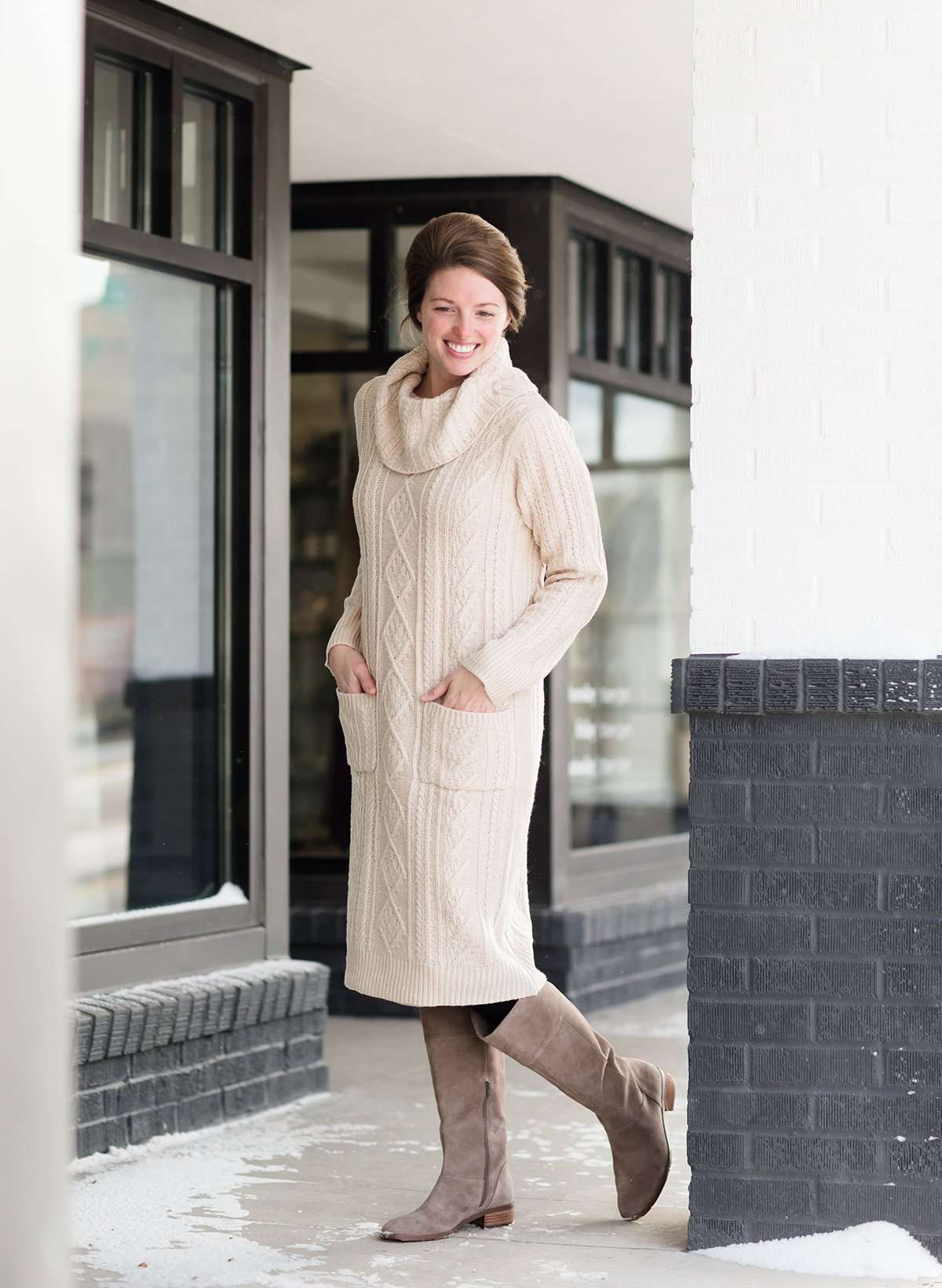 Woman wearing a cableknit, cowlneck sweater dress in taupe or burgandy. This cozy dress is below the knees and paired with tall, suede riding boots.