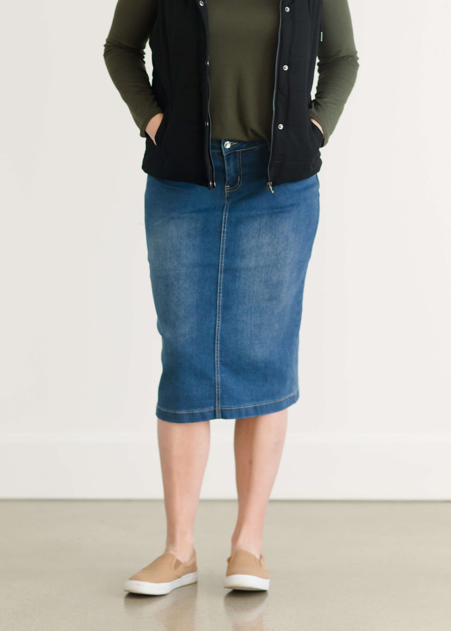 Woman wearing a mid length jean skirt that is a pencil fit. It has a jewel on the back pockets and is paired with sneakers and a olive long sleeve tee and black vest.