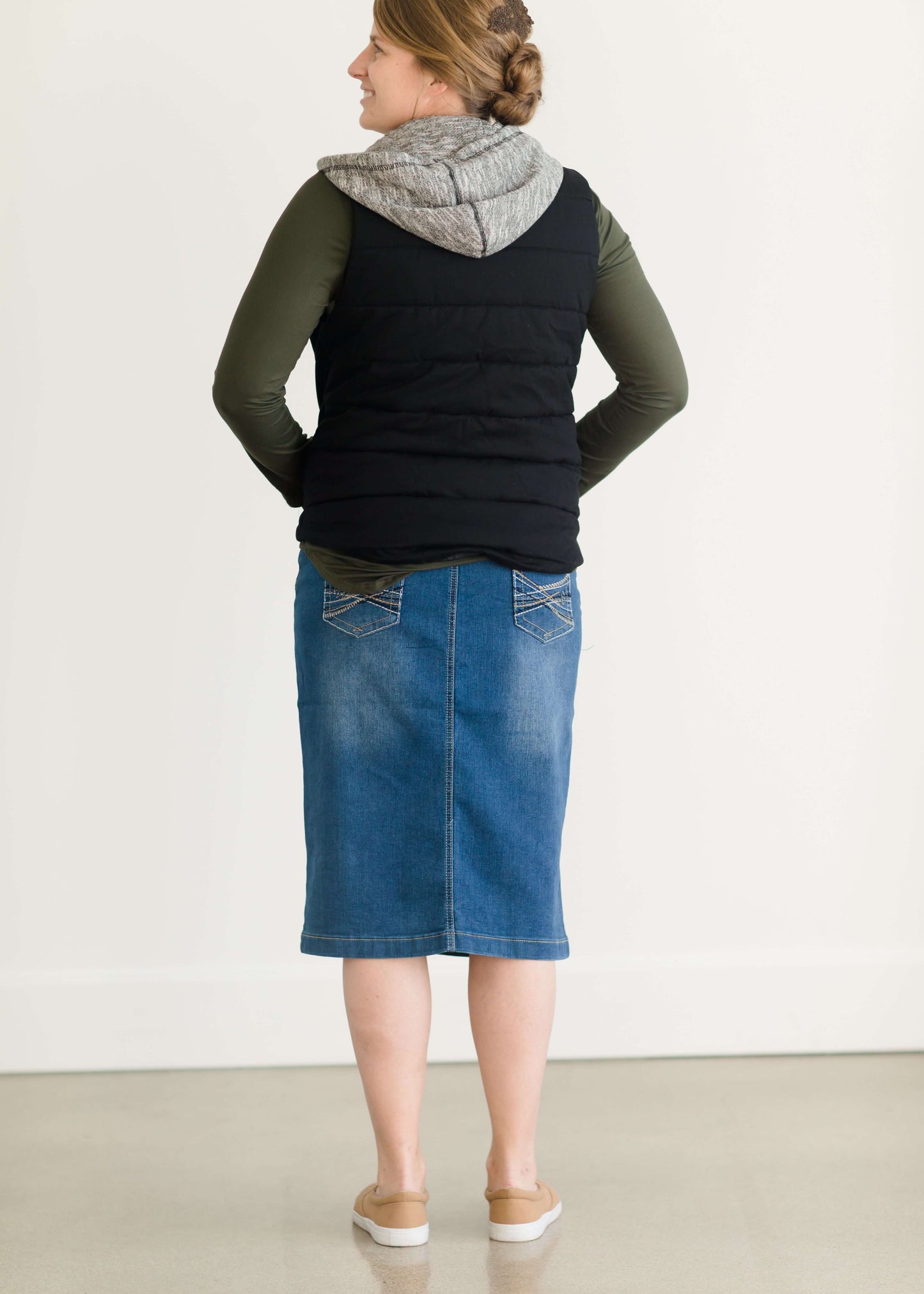 Woman wearing a mid length jean skirt that is a pencil fit. It has a jewel on the back pockets and is paired with sneakers and a olive long sleeve tee and black vest.