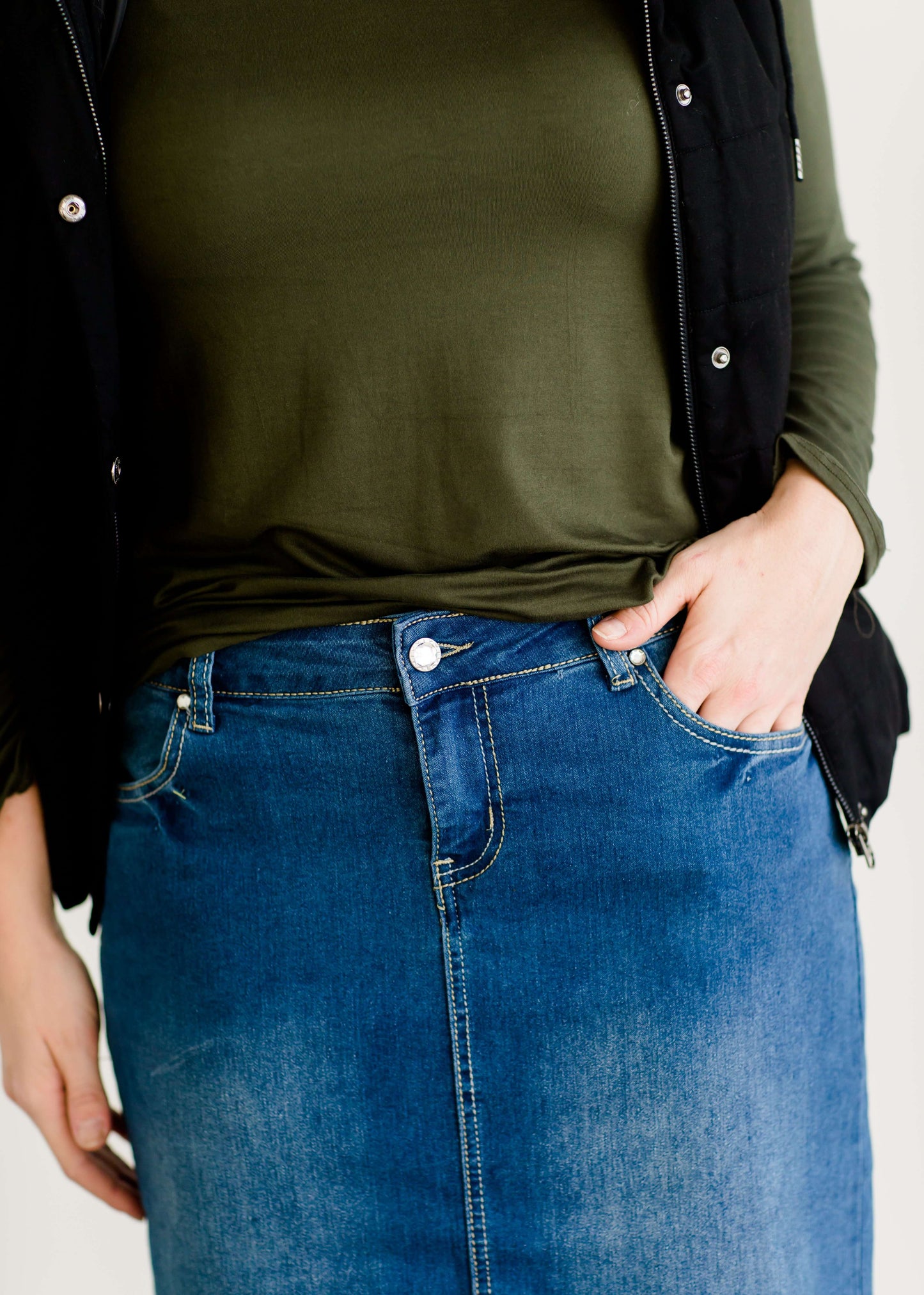 Woman wearing a mid length jean skirt that is a pencil fit. It has a jewel on the back pockets and is paired with sneakers and a olive long sleeve tee and black vest.