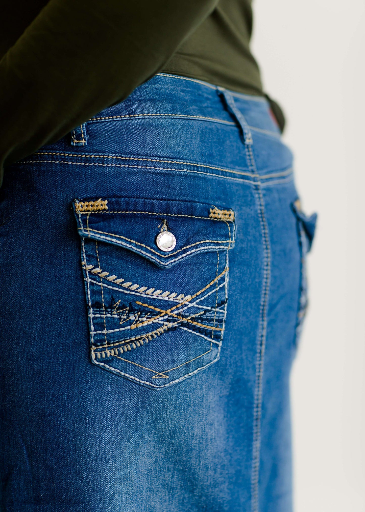 Woman wearing a mid length jean skirt that is a pencil fit. It has a jewel on the back pockets and is paired with sneakers and a olive long sleeve tee and black vest.
