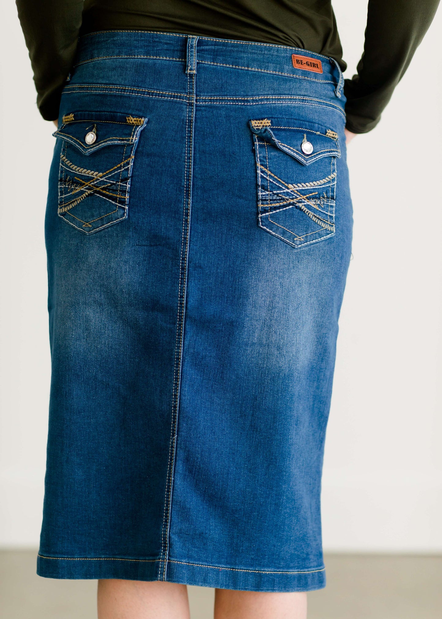 Woman wearing a mid length jean skirt that is a pencil fit. It has a jewel on the back pockets and is paired with sneakers and a olive long sleeve tee and black vest.