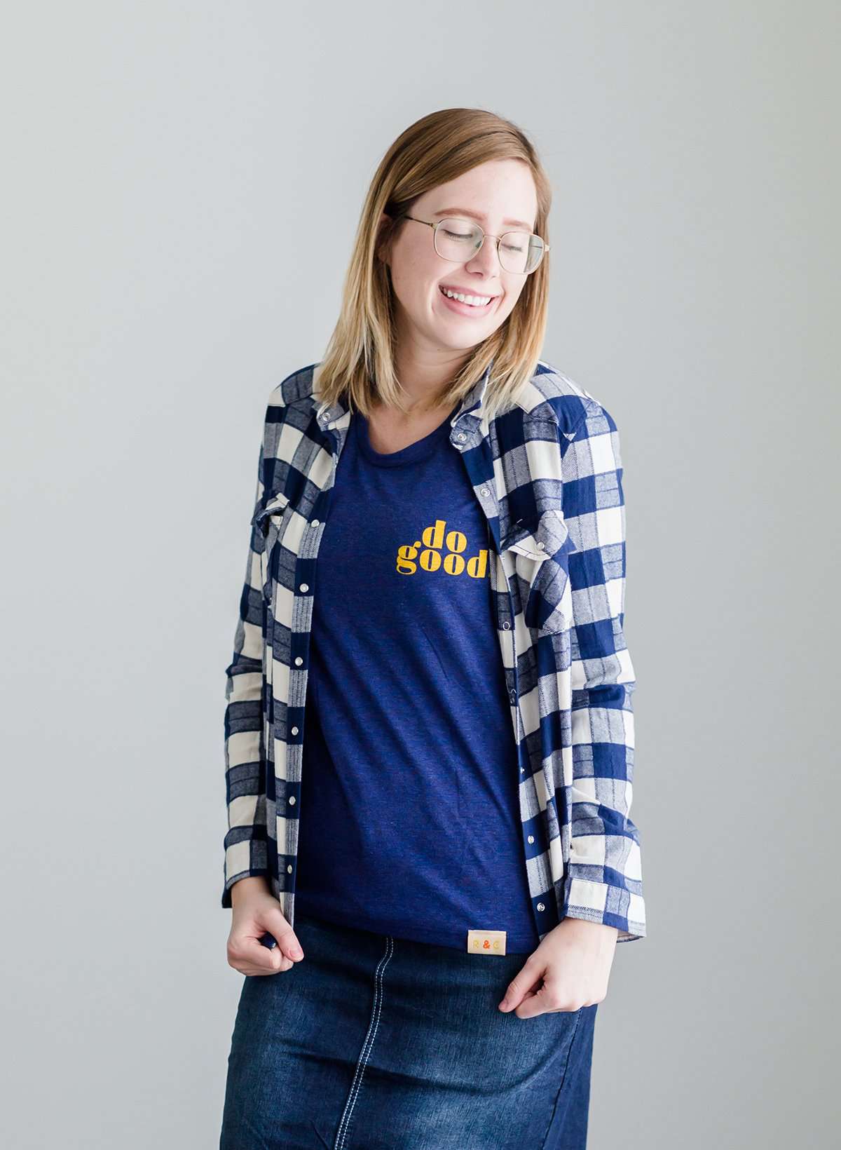 Young woman wearing a blue graphic tee with yellow words that says "Do Good." It is also paired with a below the knee denim skirt and a plaid button up top.