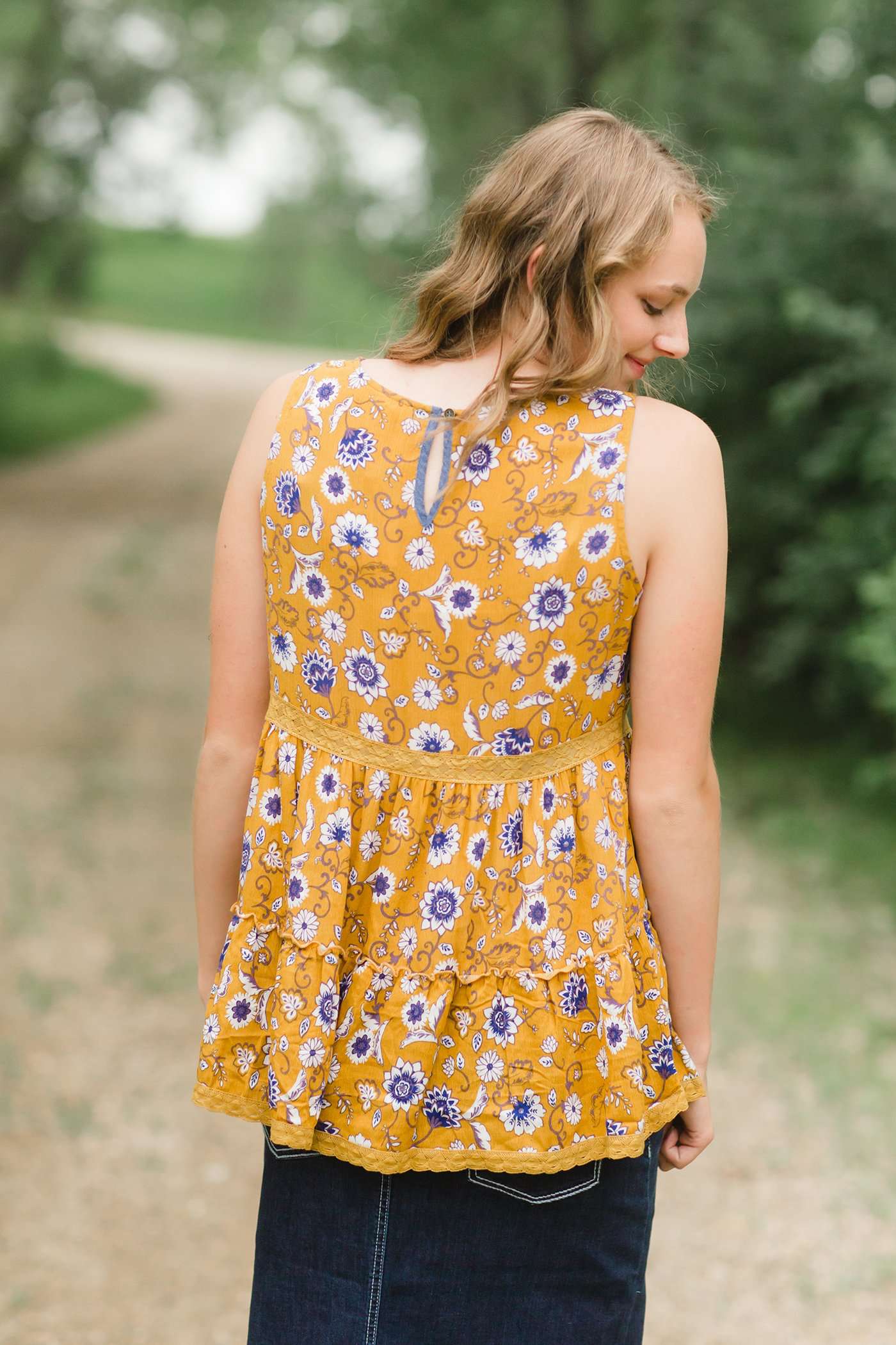 young woman wearing a mustard color tank with blue and white florals. There is embroidered detail just below the chest.