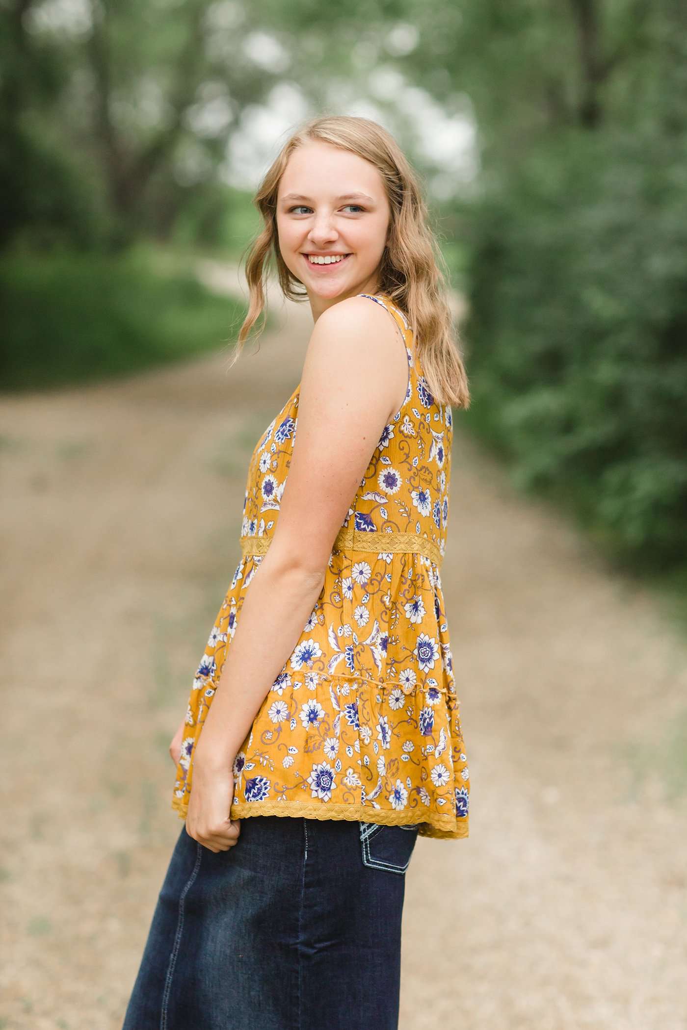 young woman wearing a mustard color tank with blue and white florals. There is embroidered detail just below the chest.