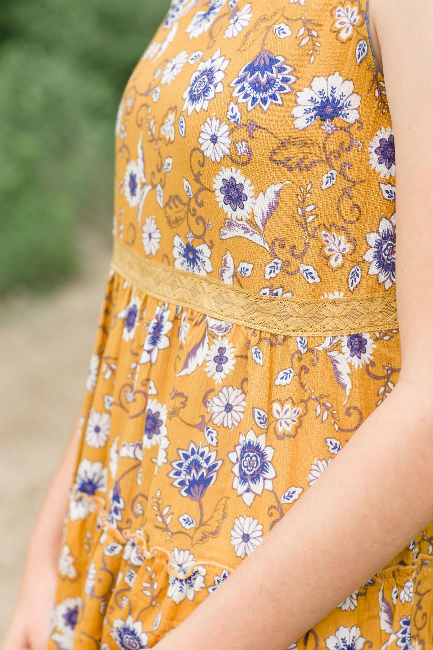 young woman wearing a mustard color tank with blue and white florals. There is embroidered detail just below the chest.