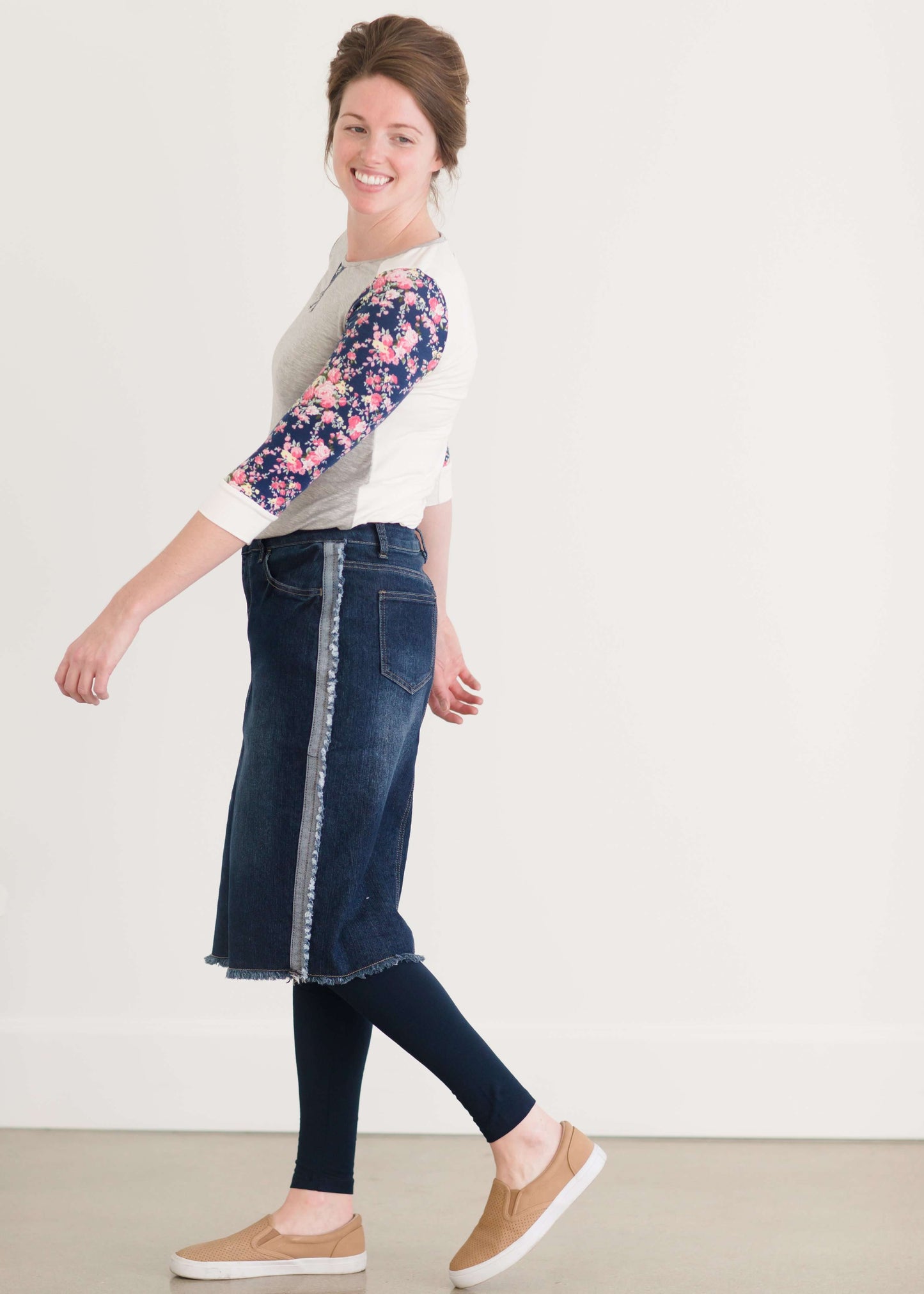 Woman wearing a below the knee jean skirt that has fringe and fray detail on the hem and side seams.