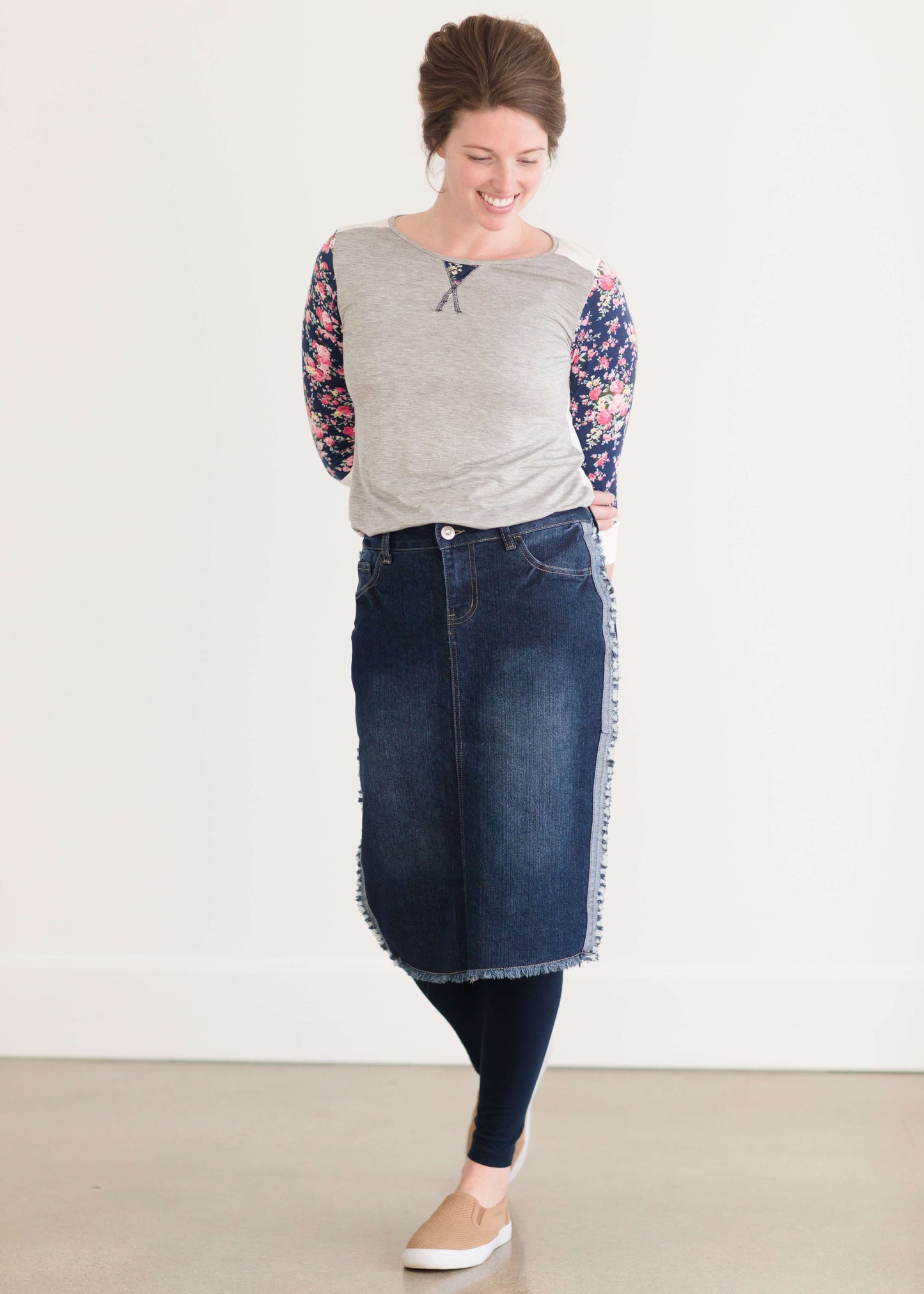 Woman wearing a below the knee jean skirt that has fringe and fray detail on the hem and side seams.