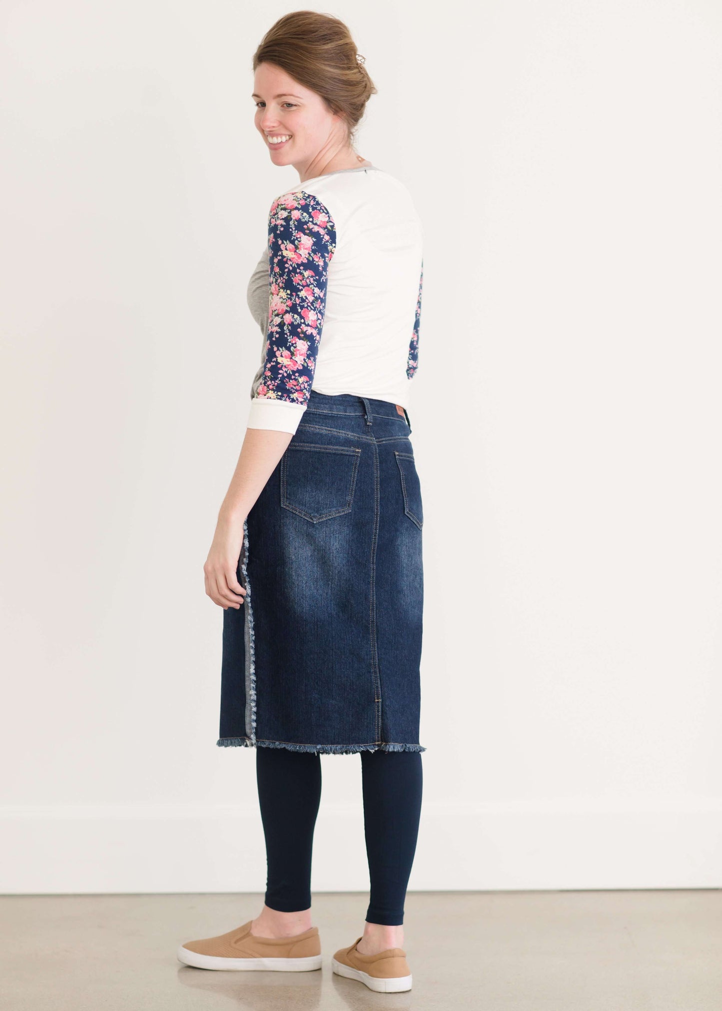 Woman wearing a below the knee jean skirt that has fringe and fray detail on the hem and side seams.