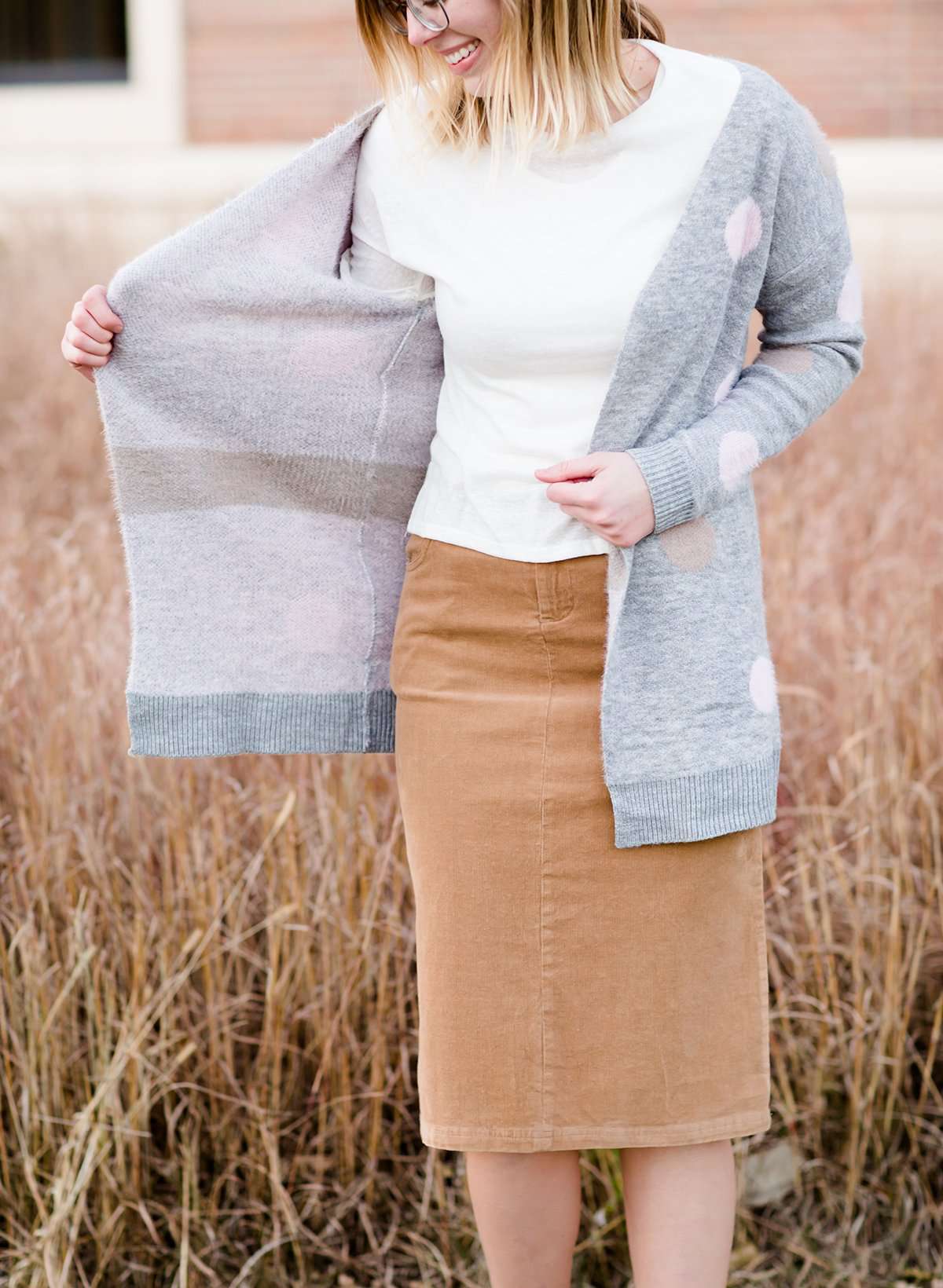 Young woman wearing a modest, long, polka dot, gray, open front cardigan. She is also wearing a khaki corduroy below the knee skirt.