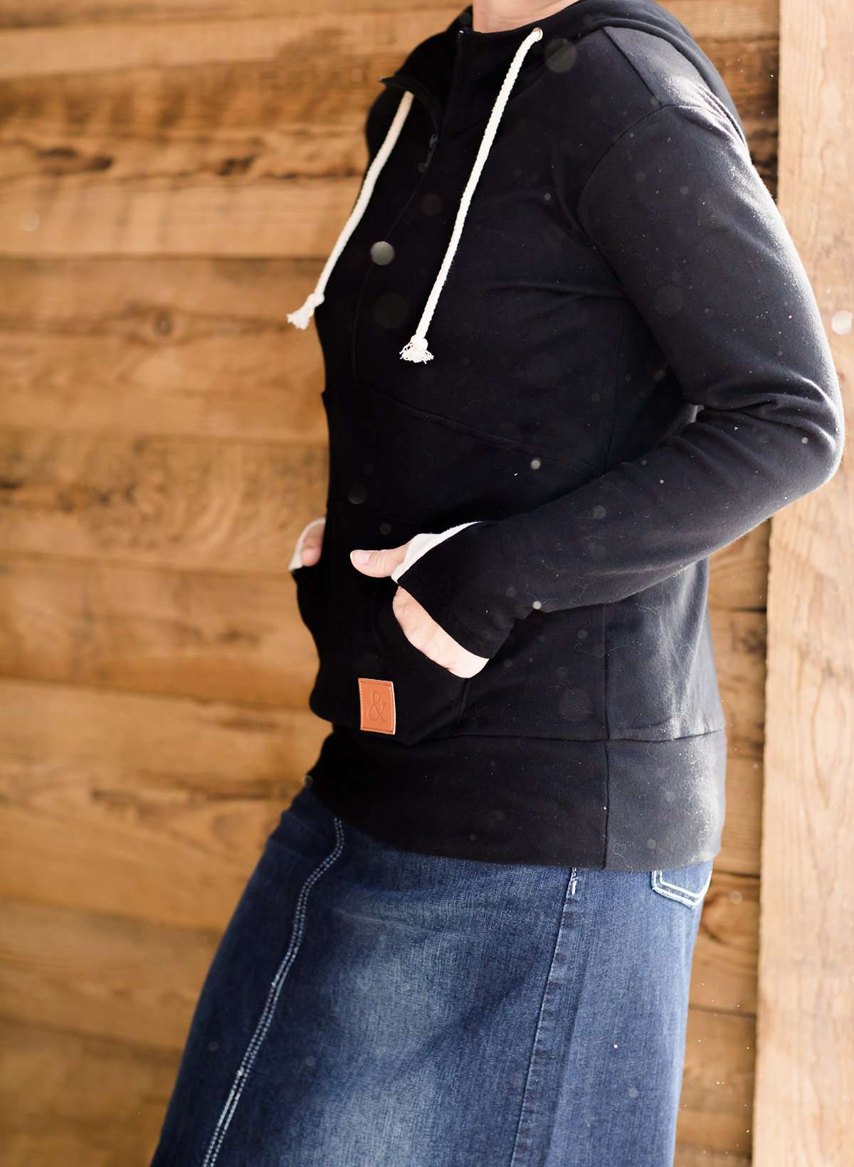Woman wearing a black, Ampersand half zip, back sweatshirt. It has white toggle strings and a front kangaroo pouch pocket. It is also paired with a long modest jean skirt.