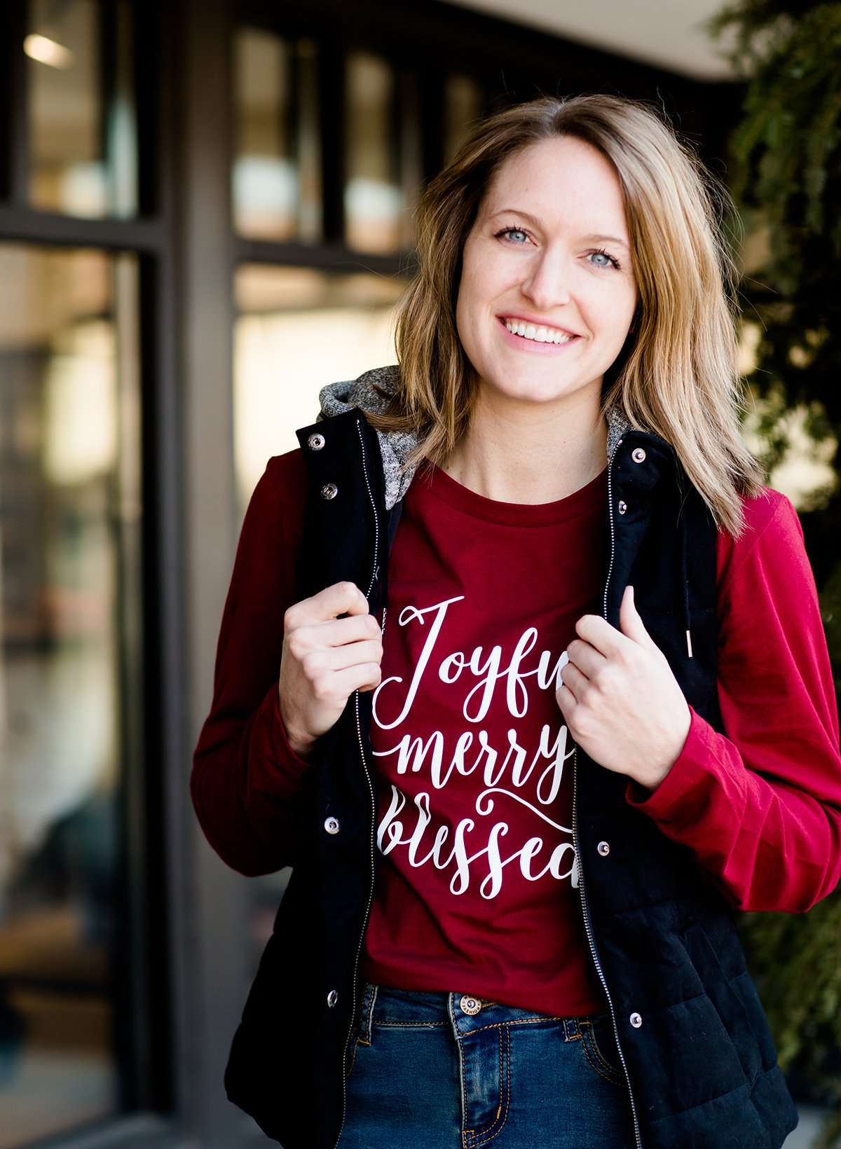 Woman standing out side of Inherit Clothing Company wearing a red and black, Joyful, Merry + Blessed Graphic Long Sleeve Tee.