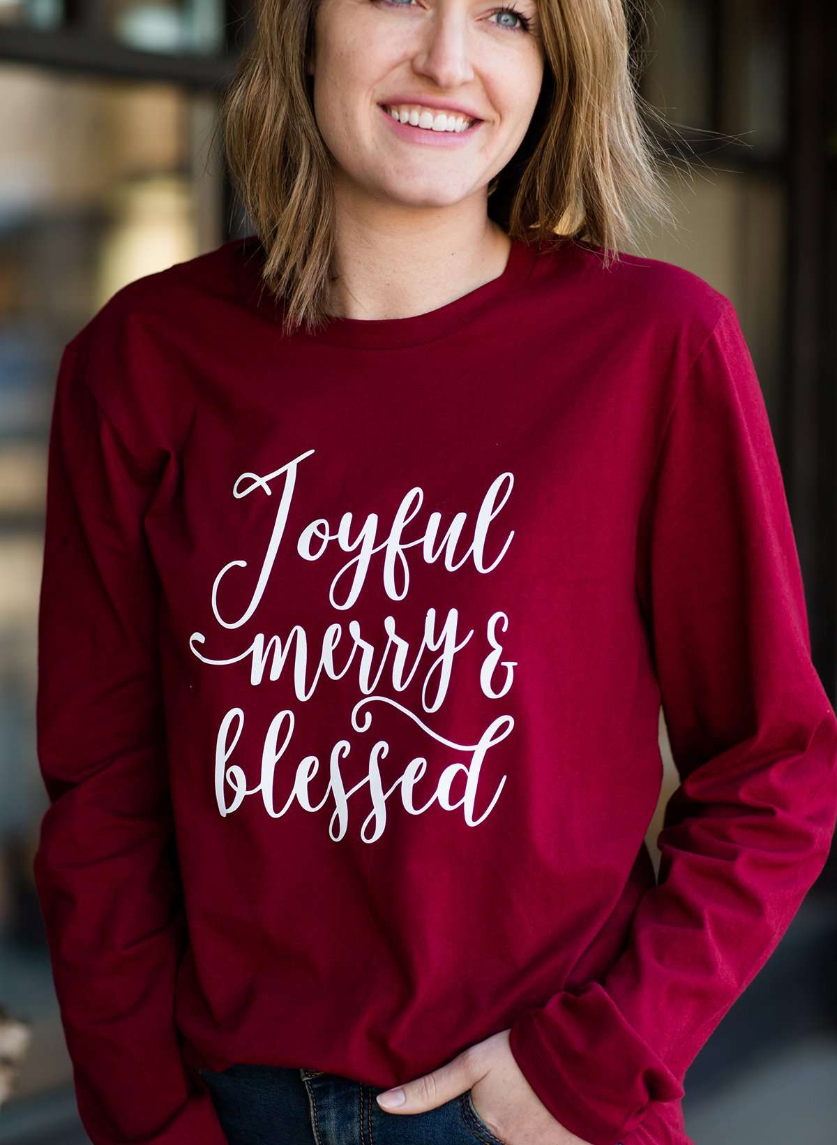 Woman standing out side of Inherit Clothing Company wearing a red and black, Joyful, Merry + Blessed Graphic Long Sleeve Tee.