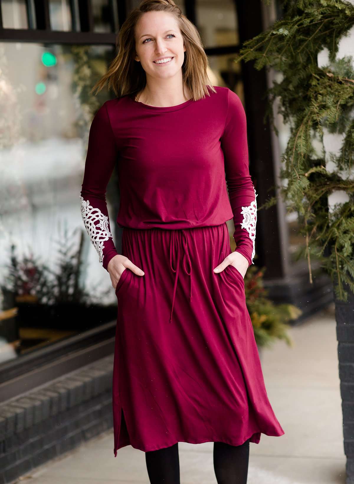 Woman wearing a burgandy and lace midi dress with black tights and sole society back boots. She is standing outside of Inherit Clothing Company.