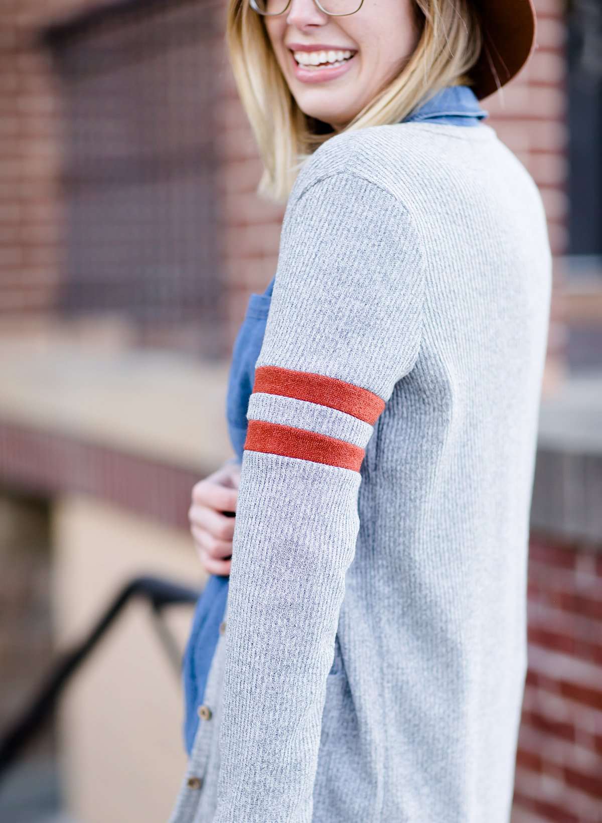 Young woman in hat and skirt wearing a gray open front long knit cardigan with orange stripes on the upper sleeve