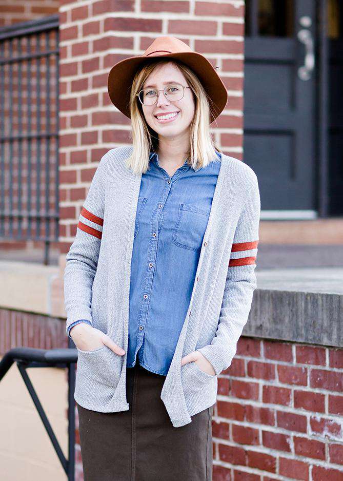Young woman in hat and skirt wearing a gray open front long knit cardigan with orange stripes on the upper sleeve