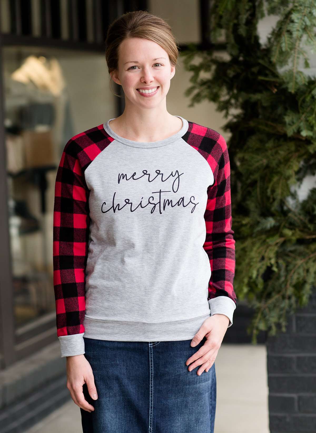 Model woman wearing a grey long sleeve tee with buffalo plaid sleeves and the script font that reads Merry Christmas.