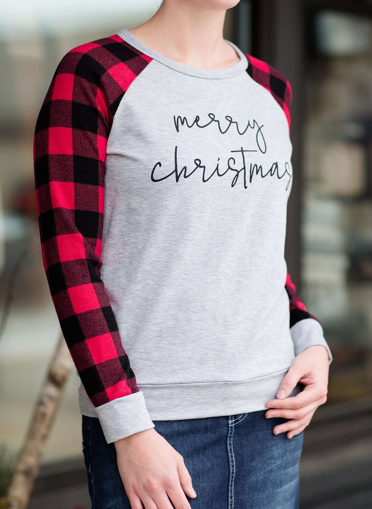 Model woman wearing a grey long sleeve tee with buffalo plaid sleeves and the script font that reads Merry Christmas.